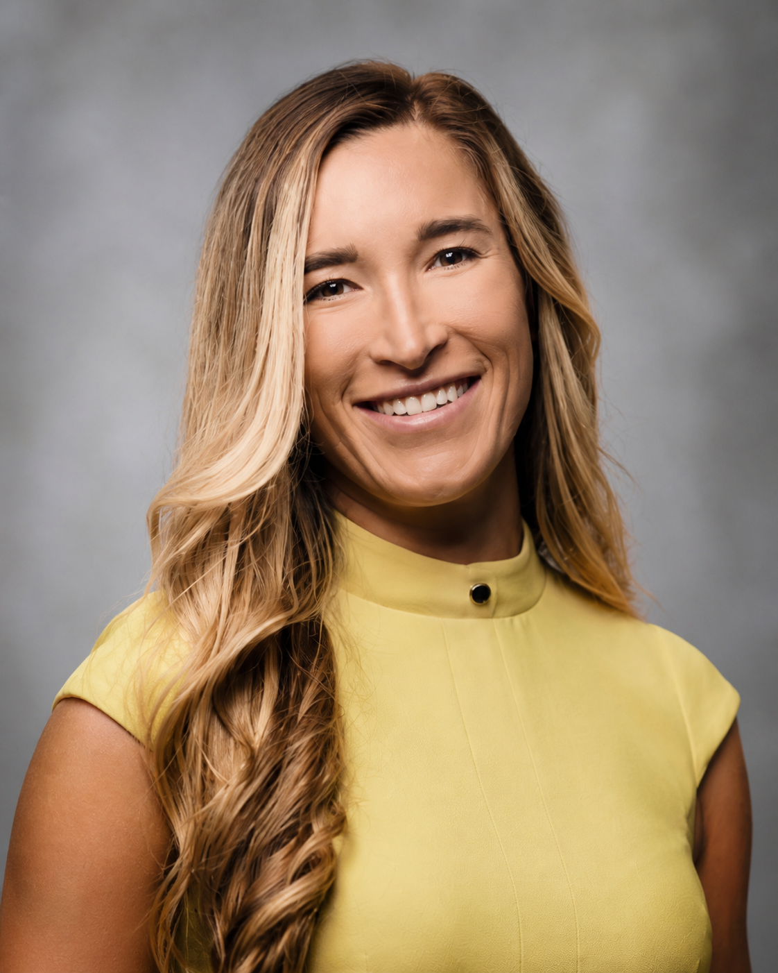 A woman with long wavy blonde hair wearing a sleeveless yellow dress with pleated hem, smiling and standing against a plain light-colored background.