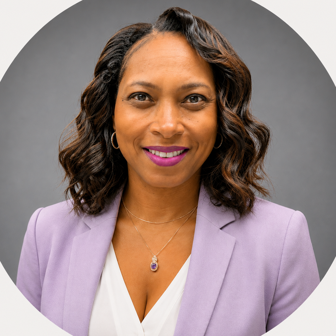A smiling African American woman with dark shoulder-length hair, wearing a blue blazer over a black top, and a delicate necklace, against a gray background.
