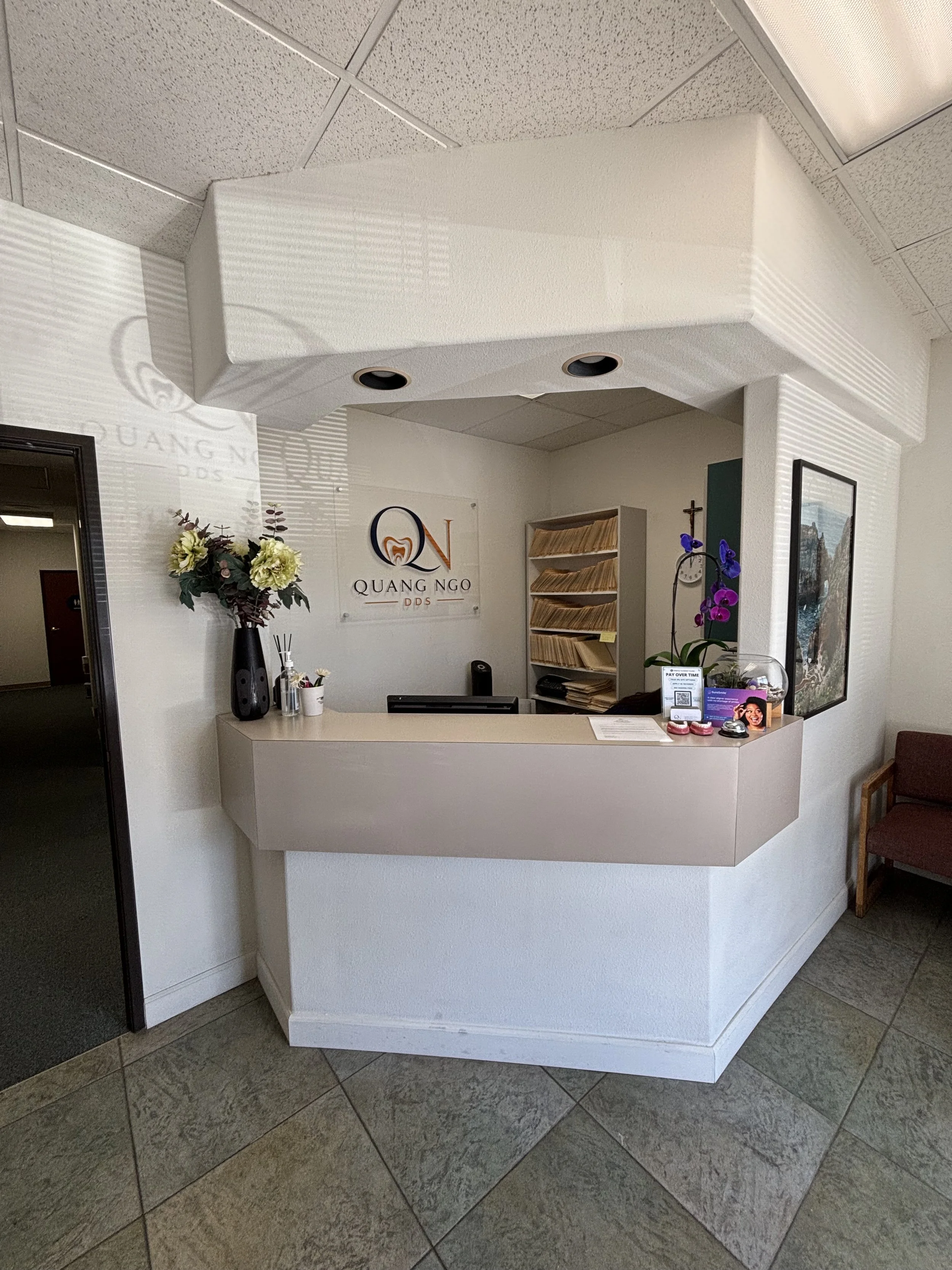 Reception area with a curved white front desk, a wall with a sign that reads 'QUANG NGO DDS', and various decorations including a flower arrangement, a framed picture, and informational pamphlets. The ceiling has a unique structure with two circular black speaker holes.