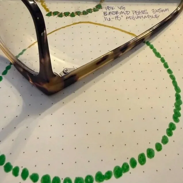Green beaded necklace design on dotted paper beside eyeglasses.