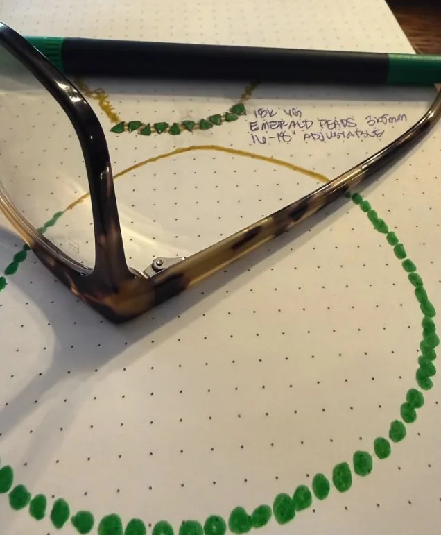 Green beaded necklace design on dotted paper beside eyeglasses.