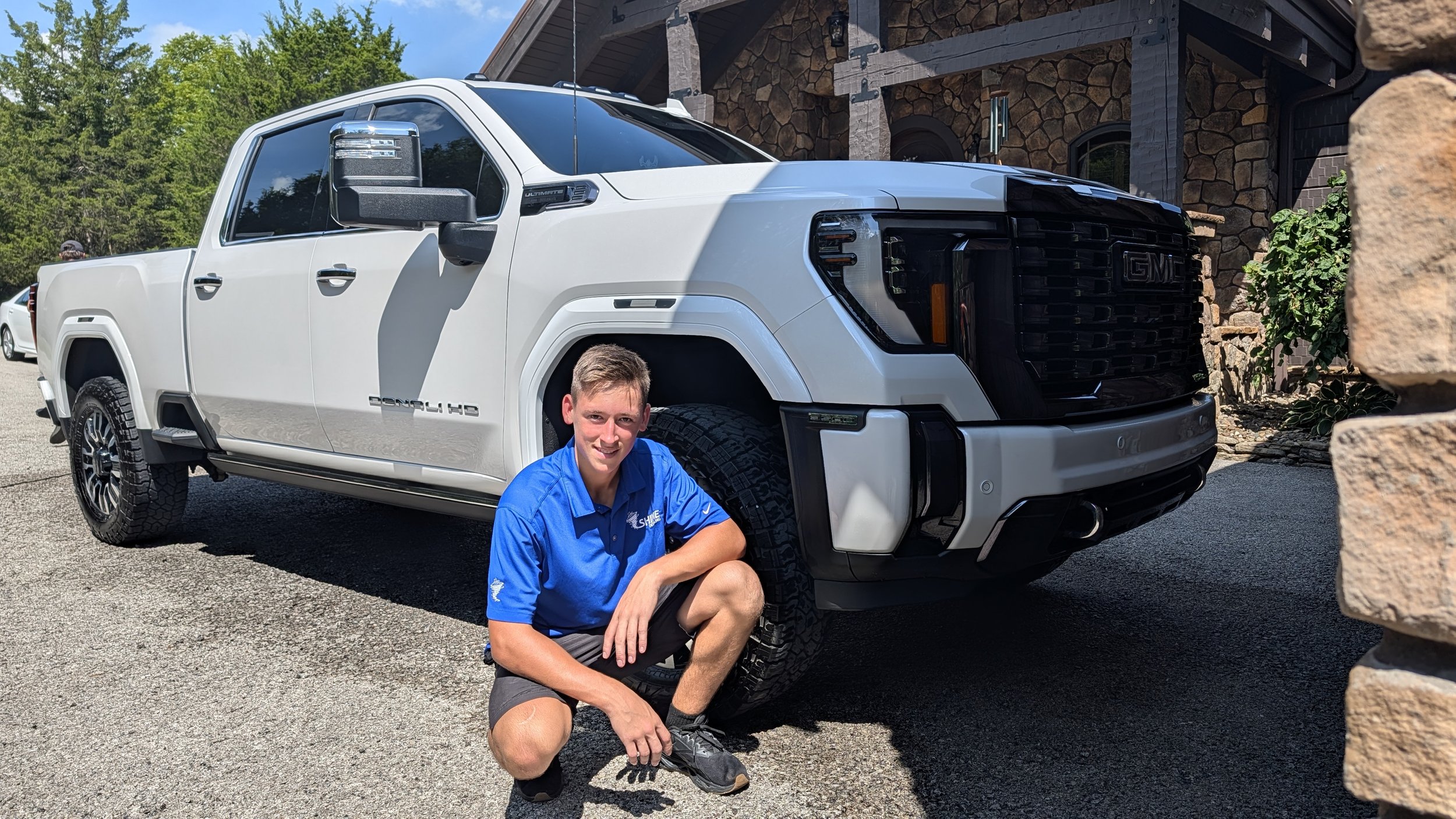 A young man in a blue shirt crouches next to a white GMC pickup truck with black trim in a driveway. The background features a stone house, trees, and a clear sky.