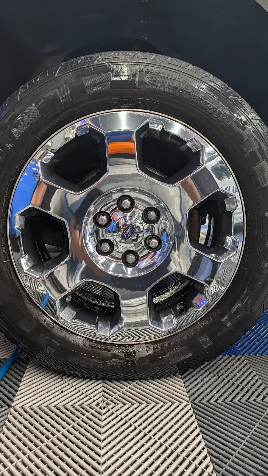 A close-up of a chrome Ford tire rim with black tire, mounted on a vehicle. The surface is shiny and reflective, and the background shows a textured floor mat.