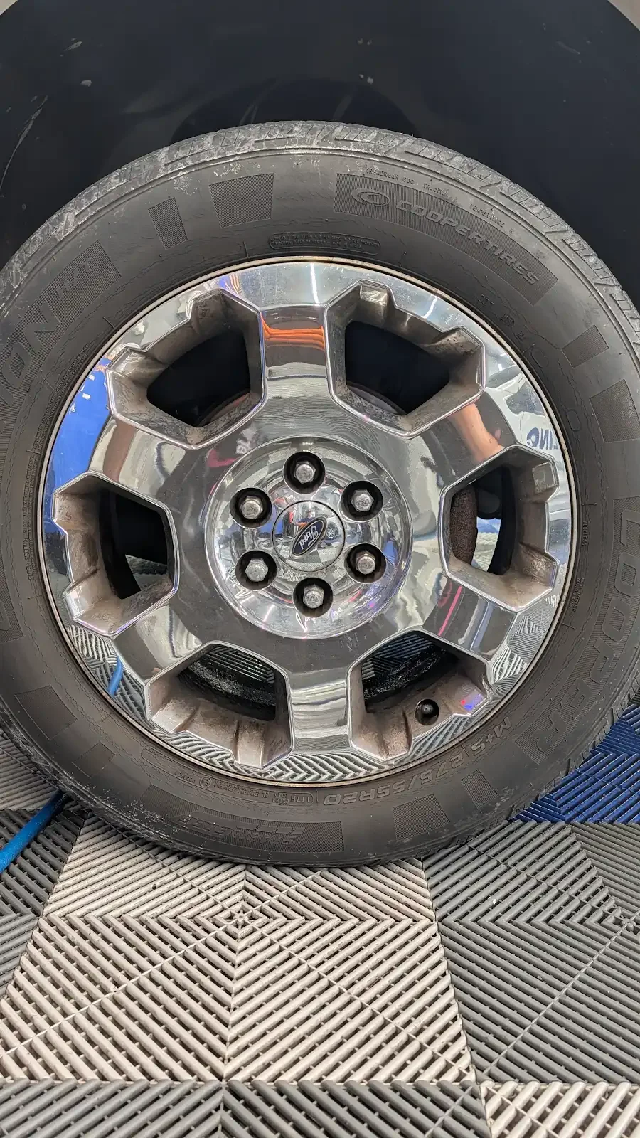 A close-up of a Ford vehicle wheel with a shiny chrome rim and a tire mounted on it, sitting on a textured surface.