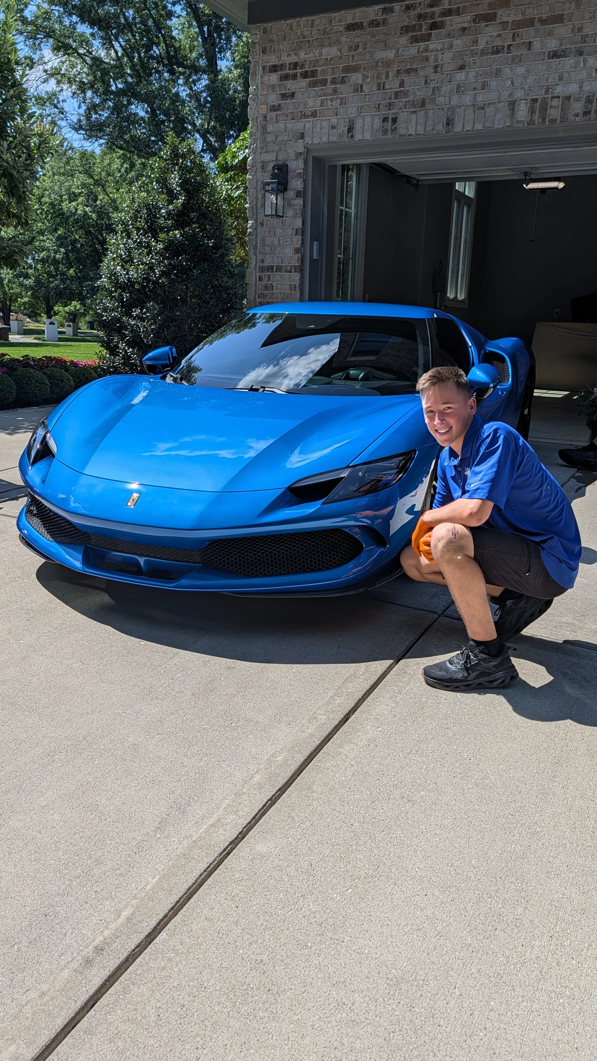 A smiling young man crouching next to a shiny blue sports car in a driveway outside a brick house.