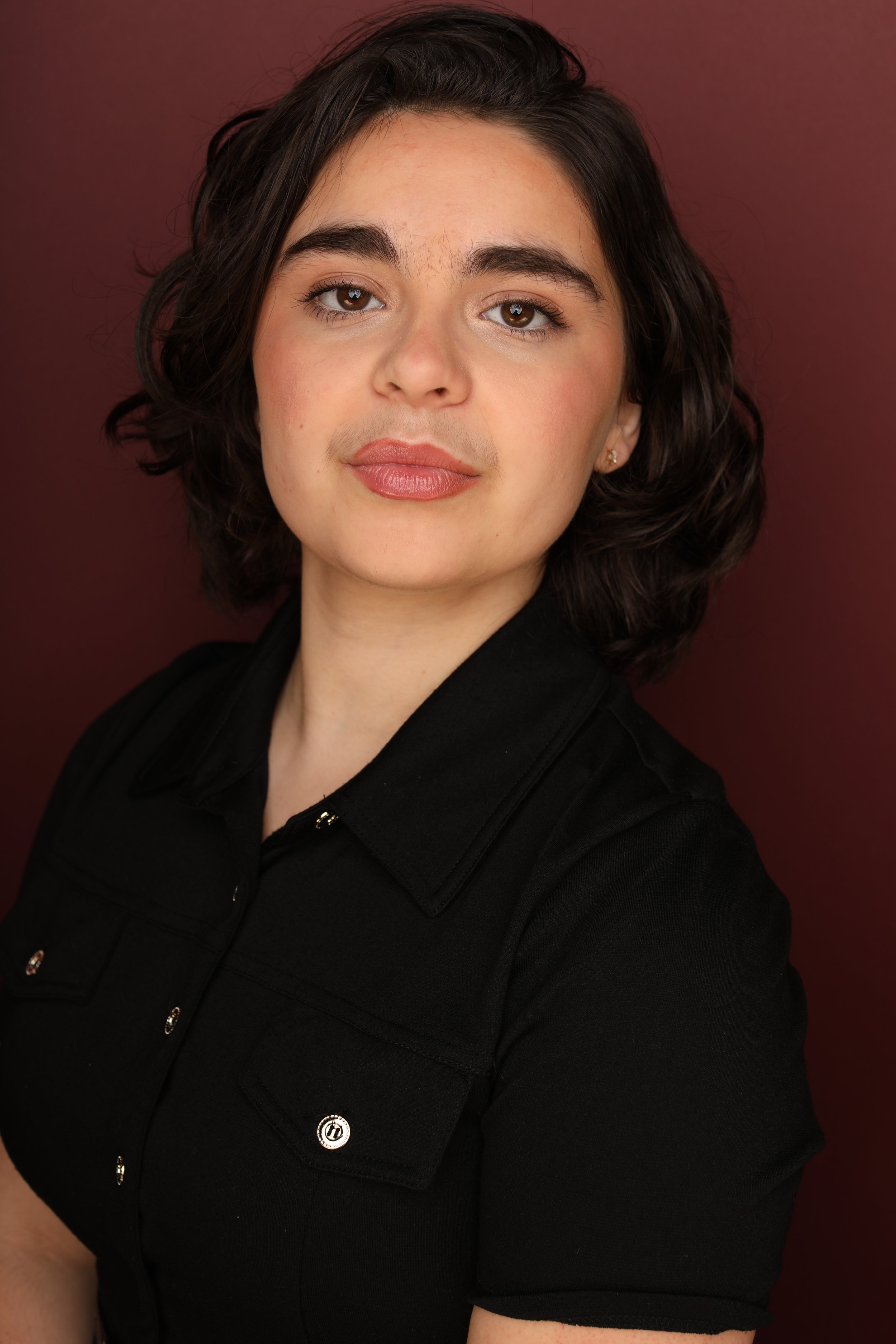 Close-up portrait of a woman with dark, wavy hair, wearing a black top, standing against a maroon background.