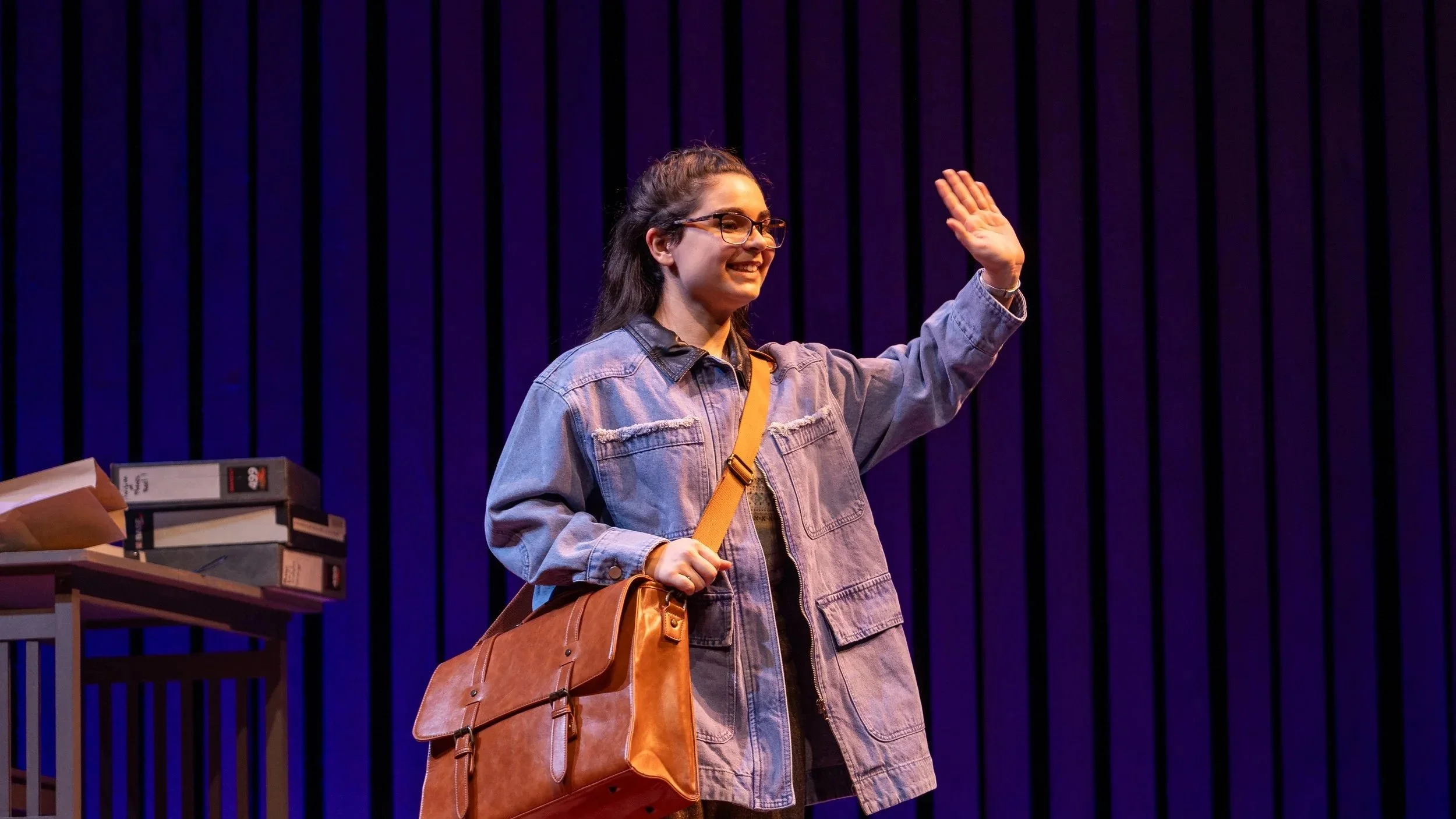 A young woman with glasses, wearing a denim jacket, smiling and waving, carrying a brown shoulder bag, standing in front of a purple striped background with a desk and stacks of books and boxes nearby.