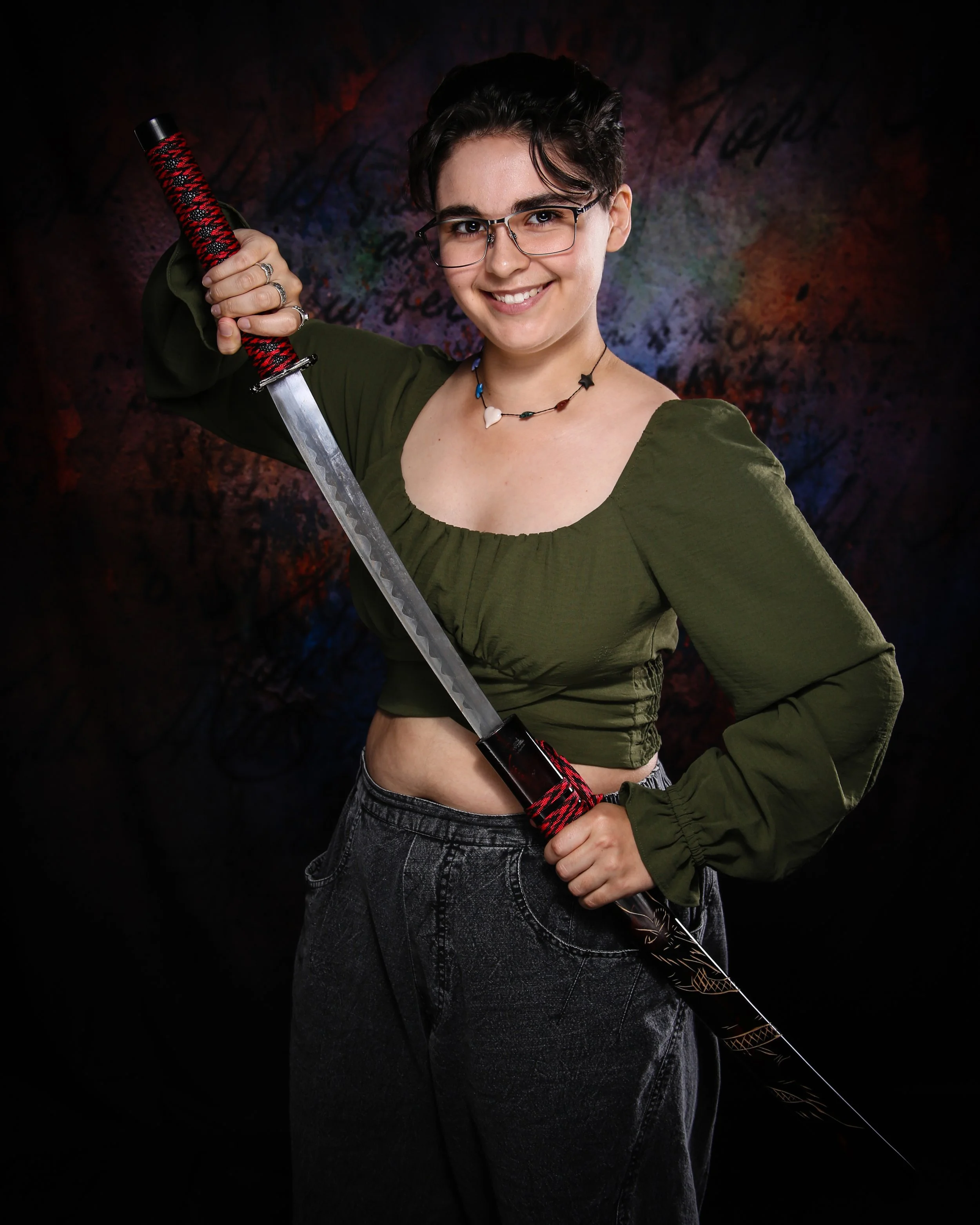 A young woman with short dark hair, glasses, and a vibrant smile, holds a decorative sword with a red and black wrapped handle, wearing a green top and black jeans, standing against a dark, colorful abstract background.