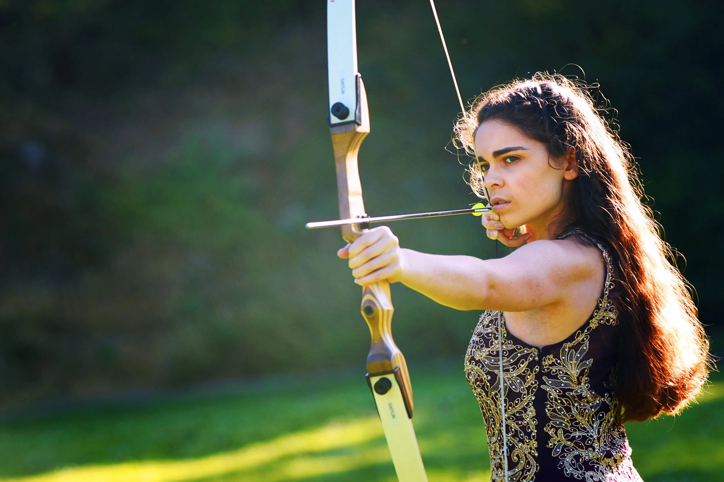 Young woman with long curly hair wearing a decorative black dress, aiming a bow and arrow outdoors with trees and greenery in the background.