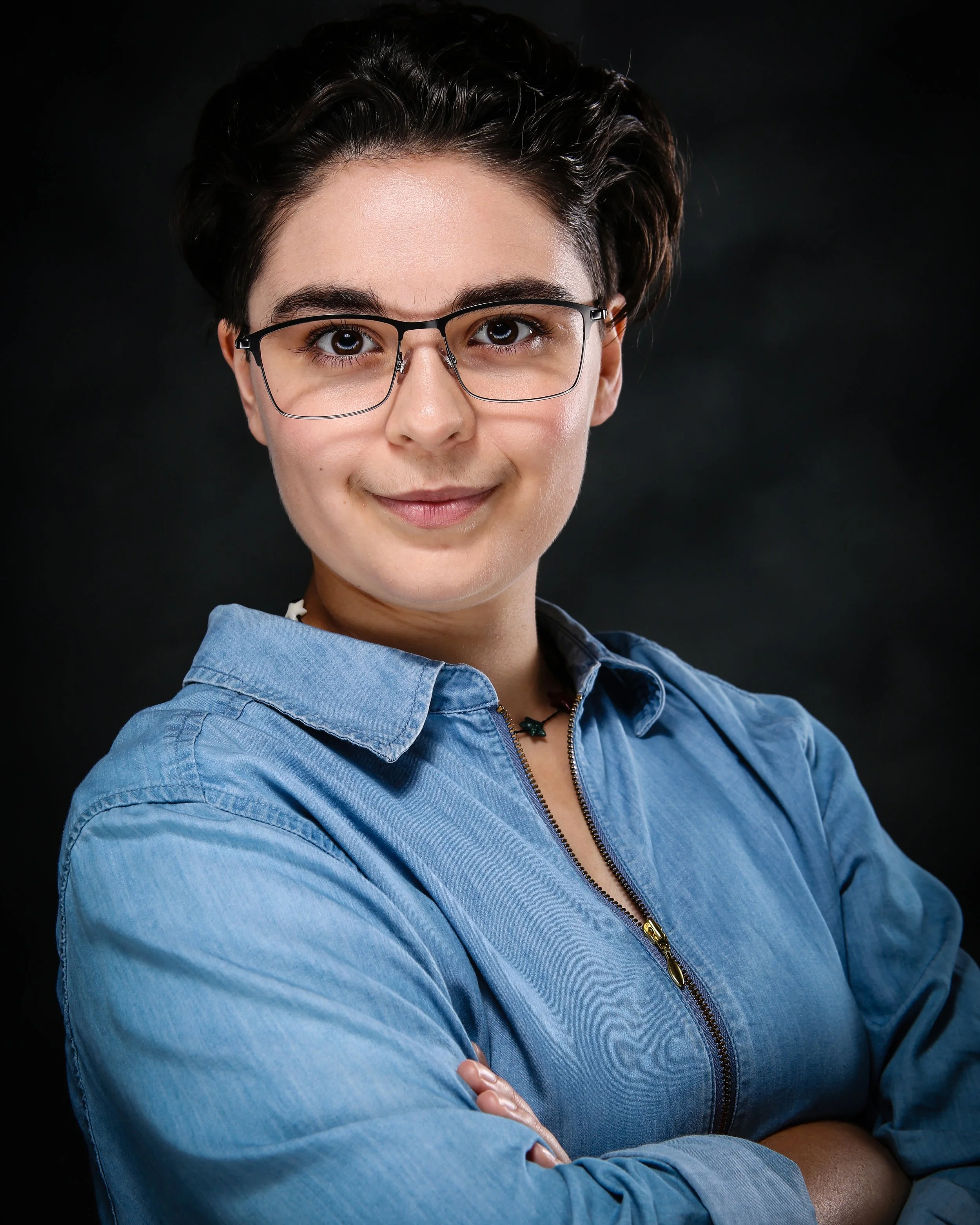 Close-up of a smiling woman with short dark hair, glasses, and a blue zip-up shirt, standing against a dark background.