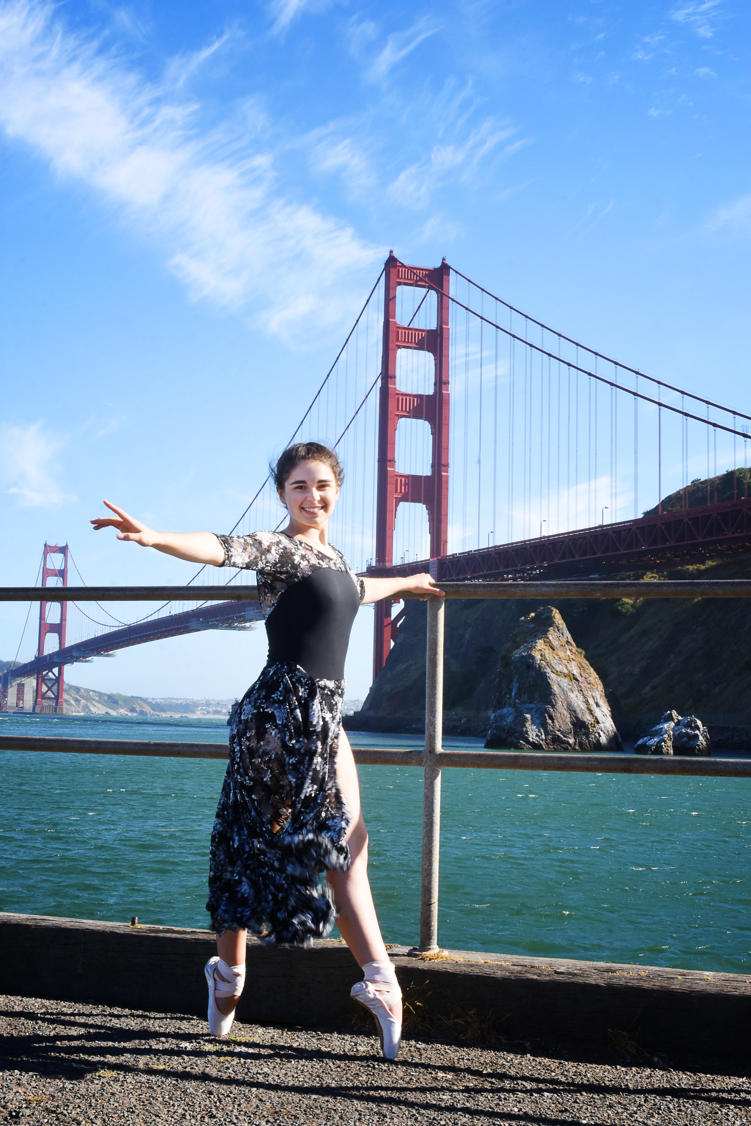 A young woman in a black and white floral dress and ballet slippers poses with arms outstretched in front of the Golden Gate Bridge in San Francisco, California, under a blue sky with wispy clouds.