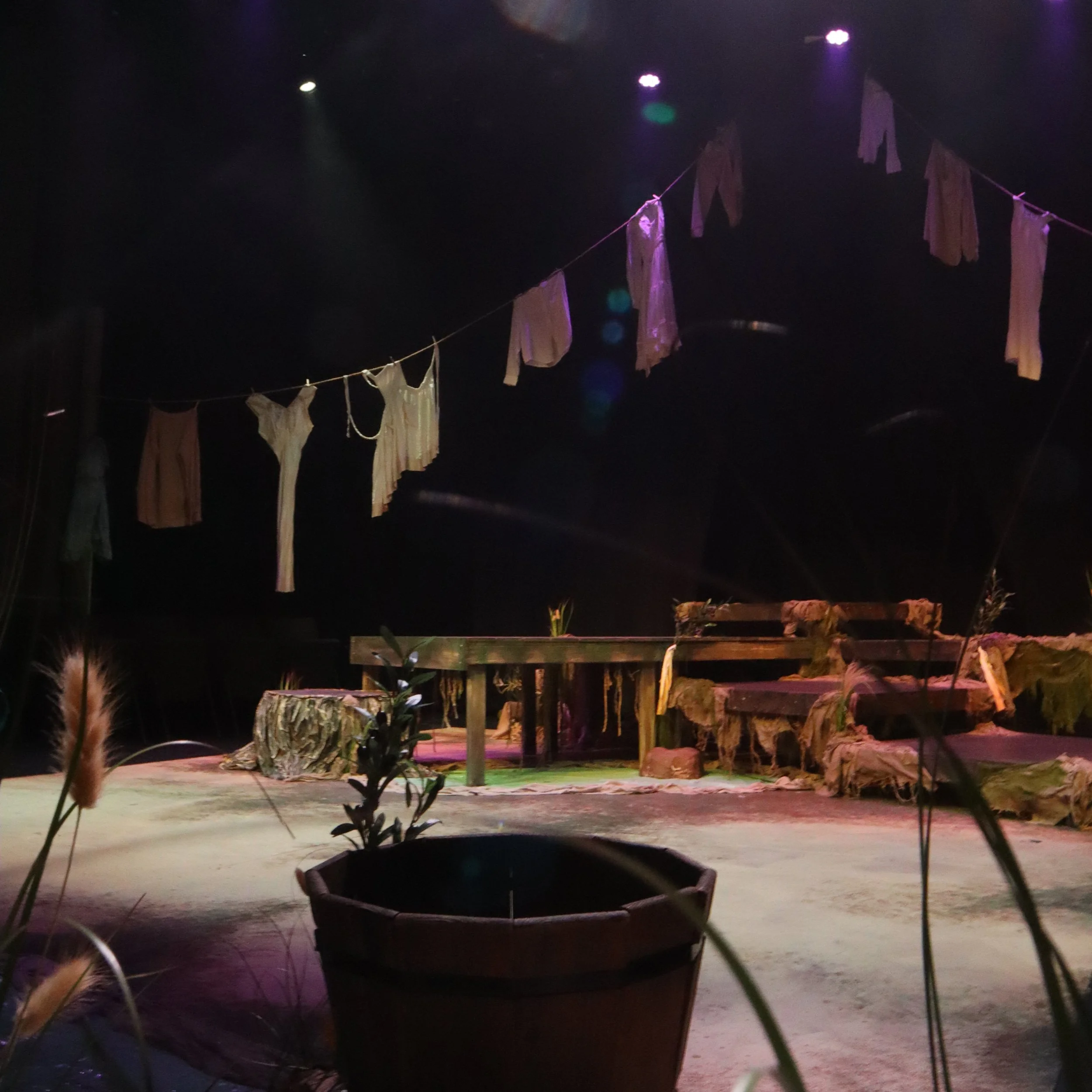 Night scene with a clothesline holding shirts and towels, illuminated by purple lighting, with plants and a wooden bench in the foreground.