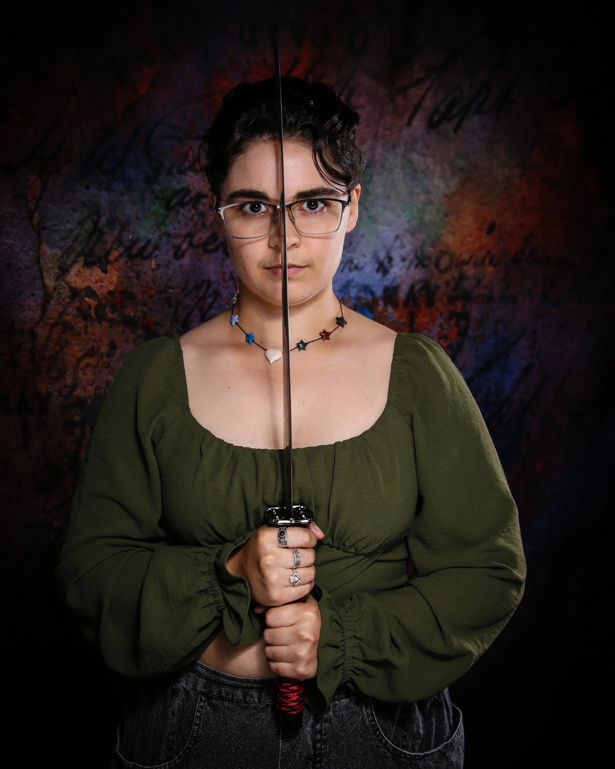 A person with short dark hair, glasses, and multiple rings on their fingers, holding a sword vertically in front of their face, wearing a green long-sleeve shirt and patterned black pants, standing against a dark, graffiti-covered wall background.