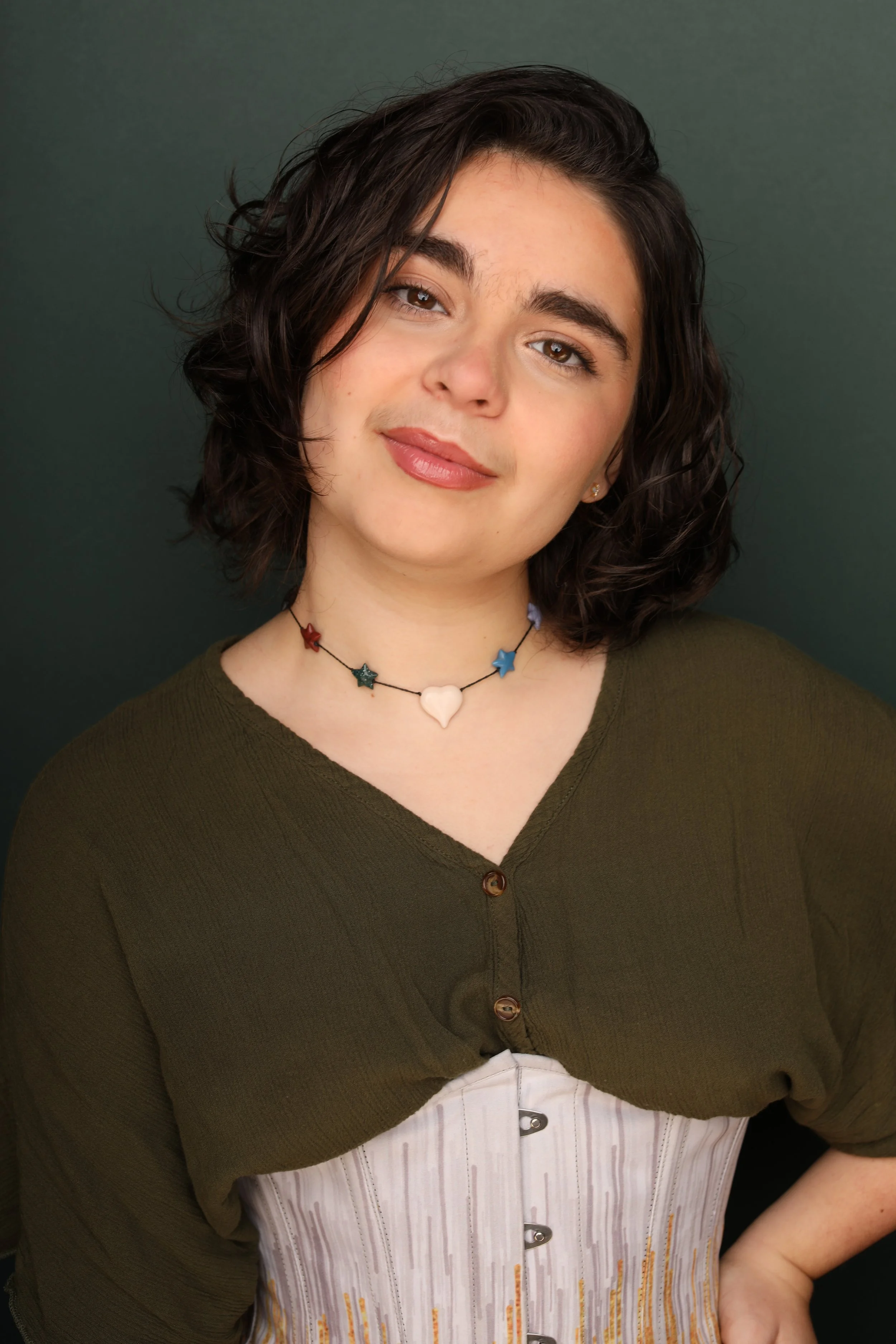 A woman with dark wavy hair, light skin, and brown eyes, wearing a green button-up top, a star and heart necklace, and earrings, smiling softly against a dark green background.