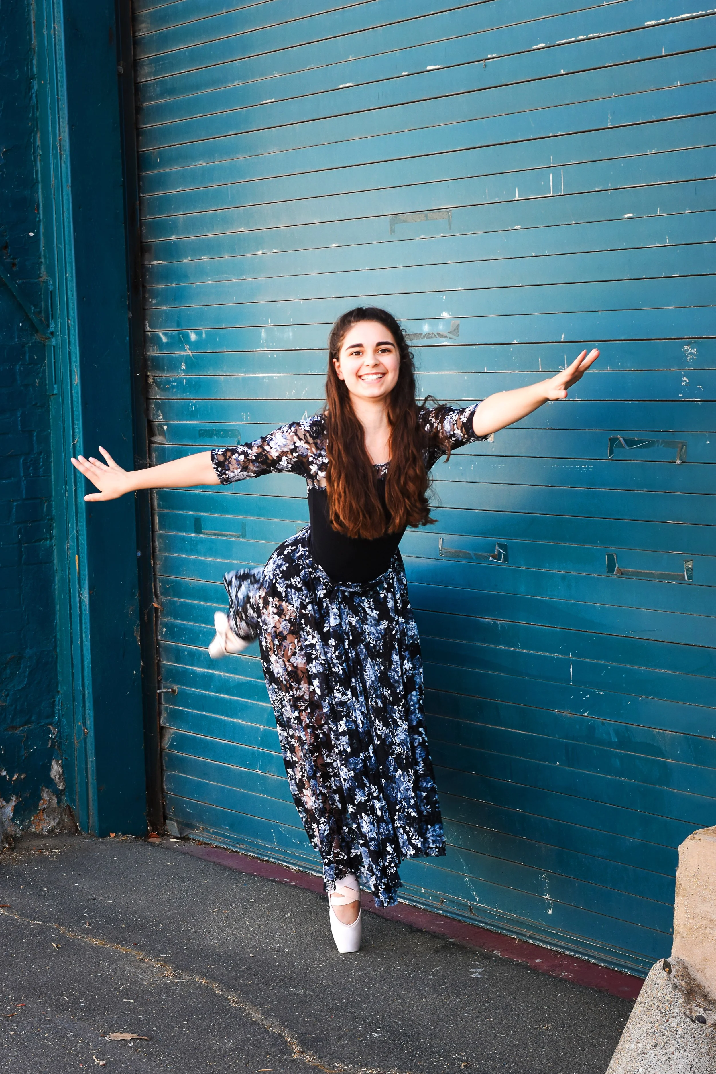 A woman with long dark hair, wearing a floral dress and white ballet pointe shoes, is standing with arms outstretched in front of a blue metal garage door. She is smiling and showing a joyful expression.