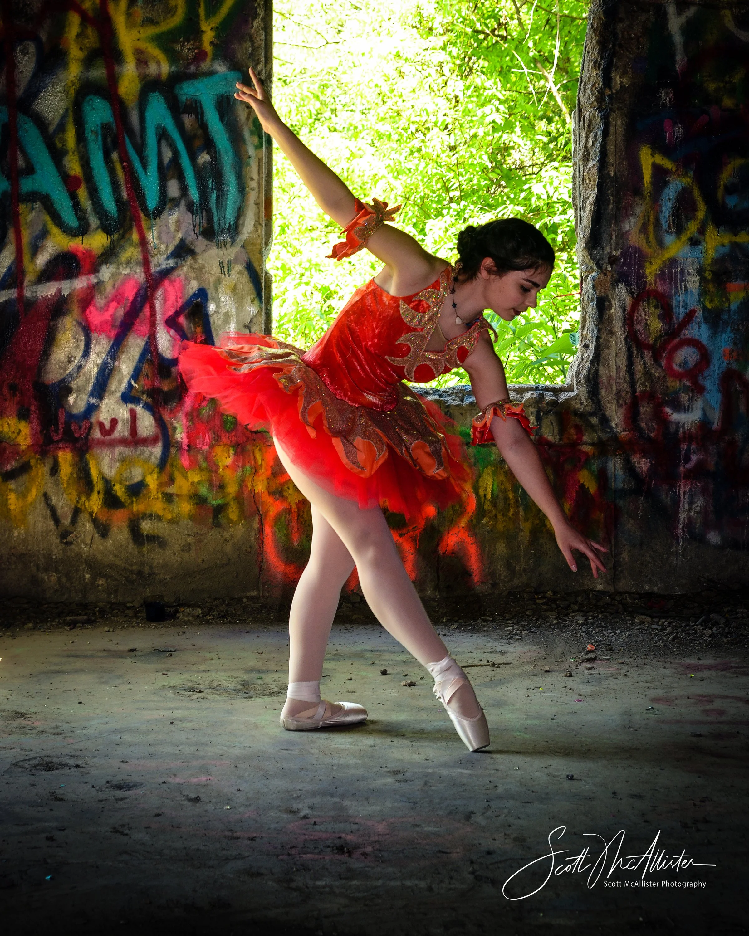 A ballerina in a red tutu and ballet shoes is practicing ballet in an abandoned building with graffiti-covered walls and a window showing green foliage outside.