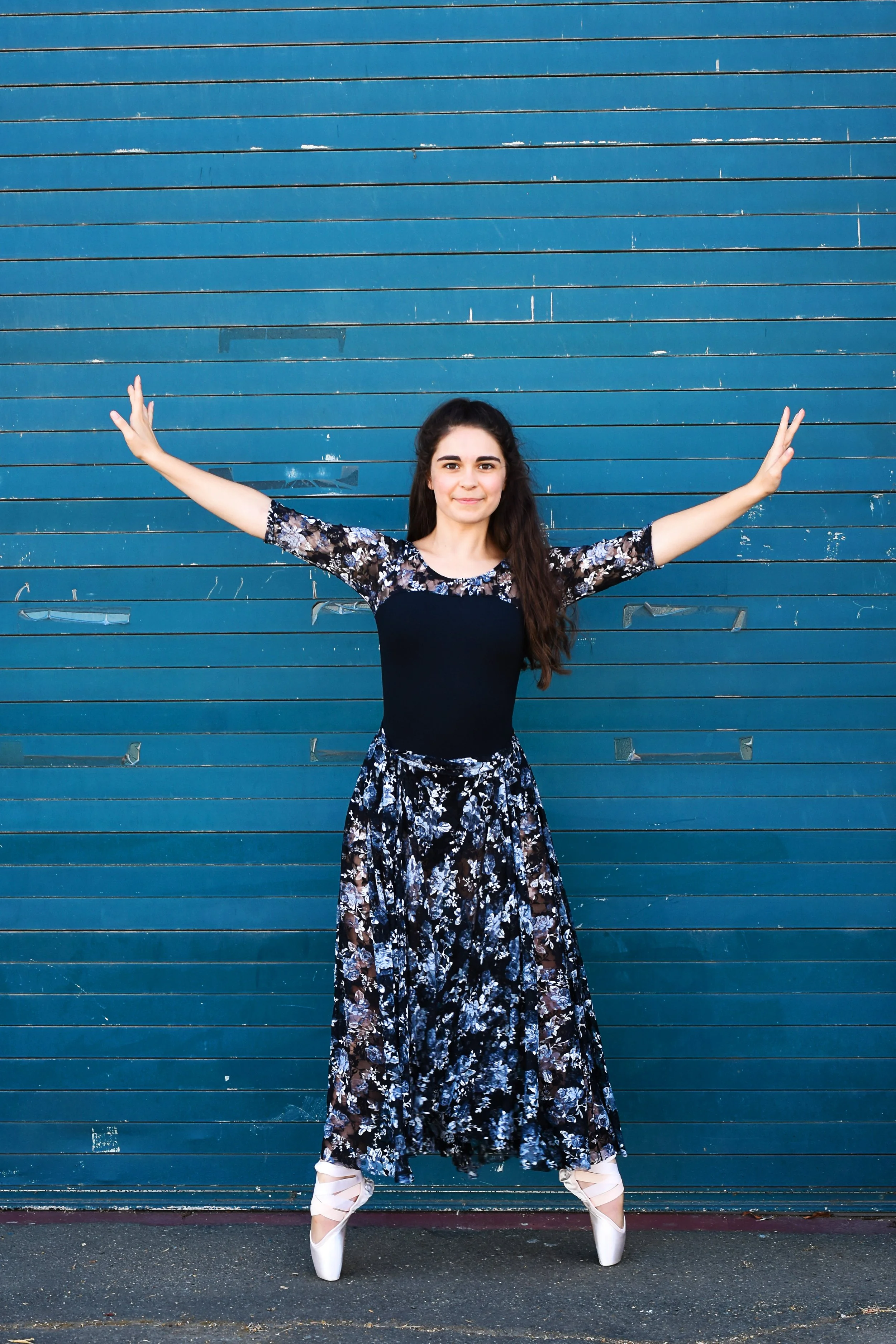 Young woman wearing a black and floral dress with ballet pointe shoes, standing with arms stretched out against a blue garage door with peeling paint.