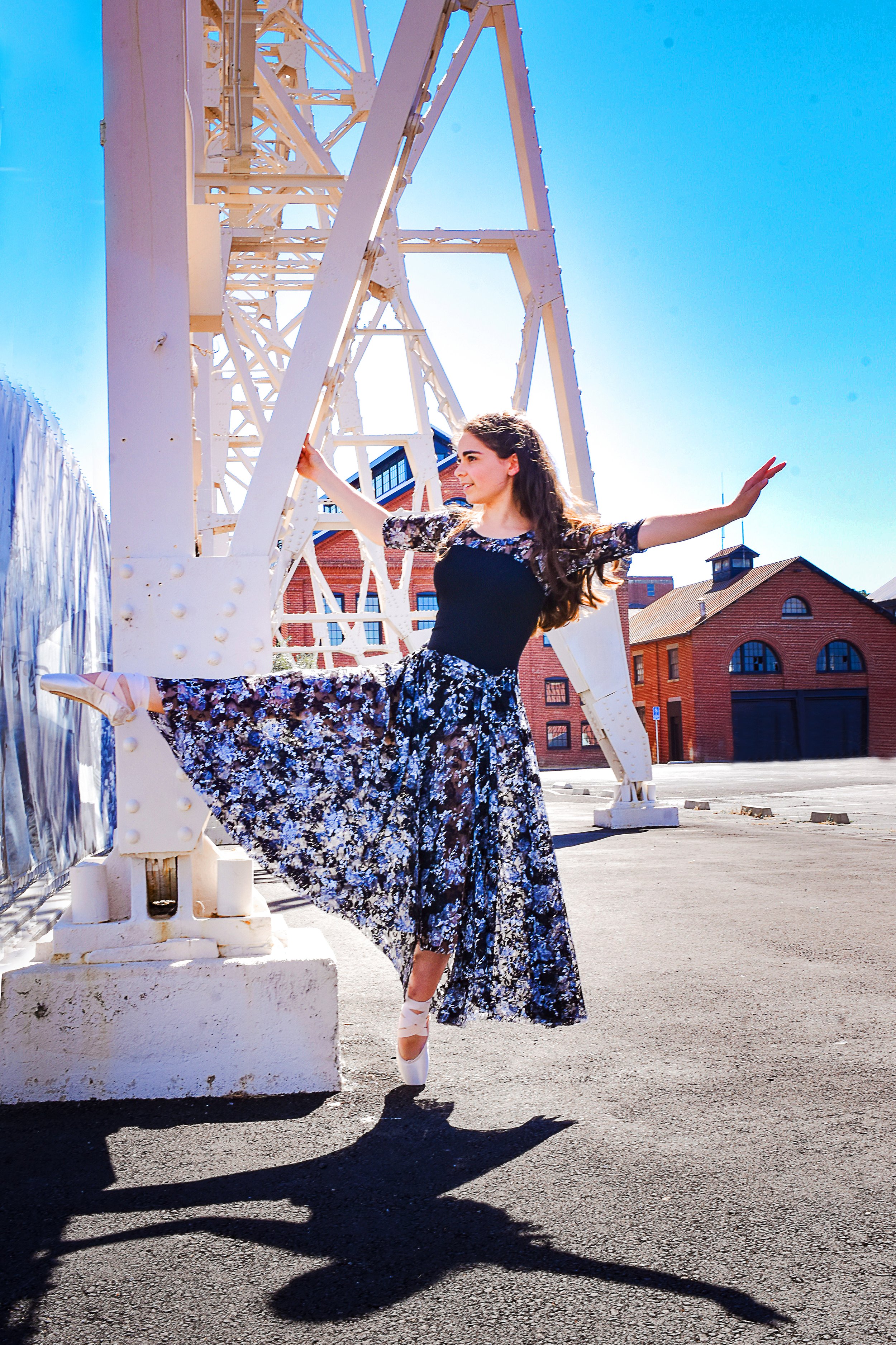 A young woman in a black and floral dress poses with her leg raised and arms extended in front of a white industrial structure on a sunny day, with brick buildings in the background.