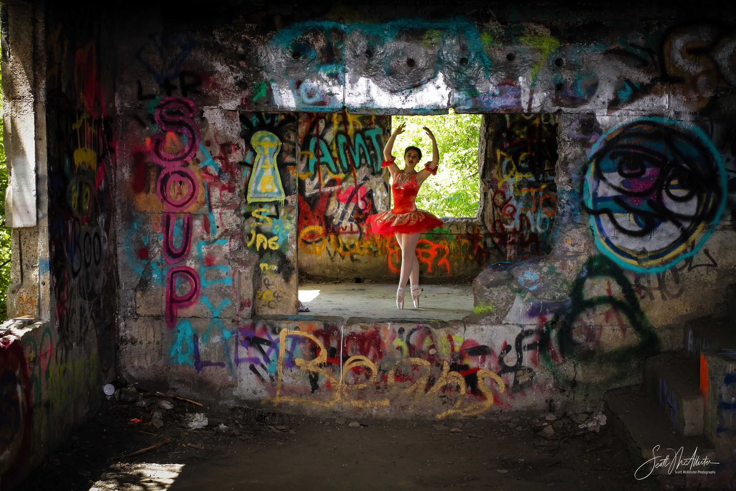 A ballerina in a red tutu practicing ballet inside an abandoned building with graffiti-covered walls and a broken window.