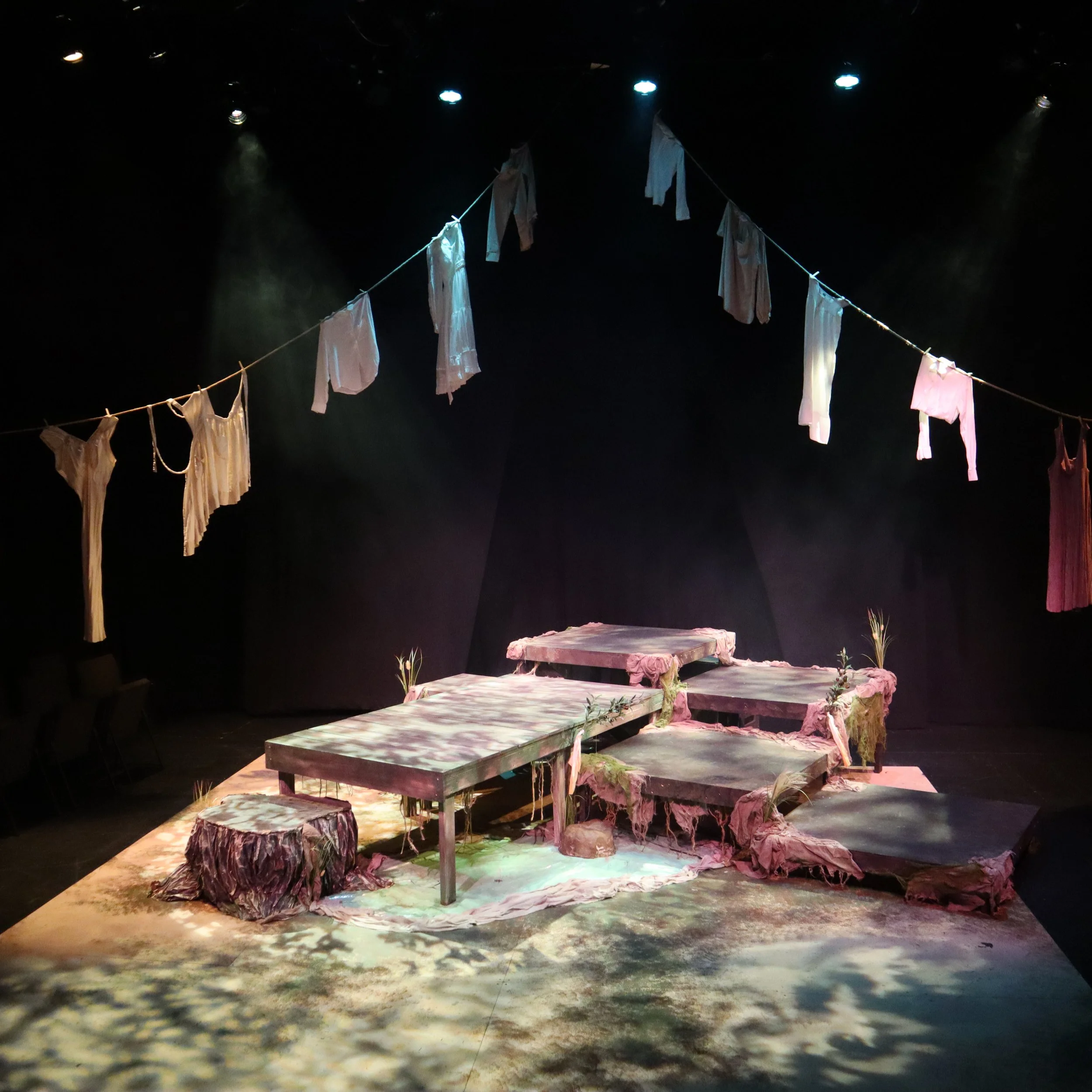 Stage set with wooden platforms and fabric resembling clouds, with hanging laundry on a line and small plants on stage.