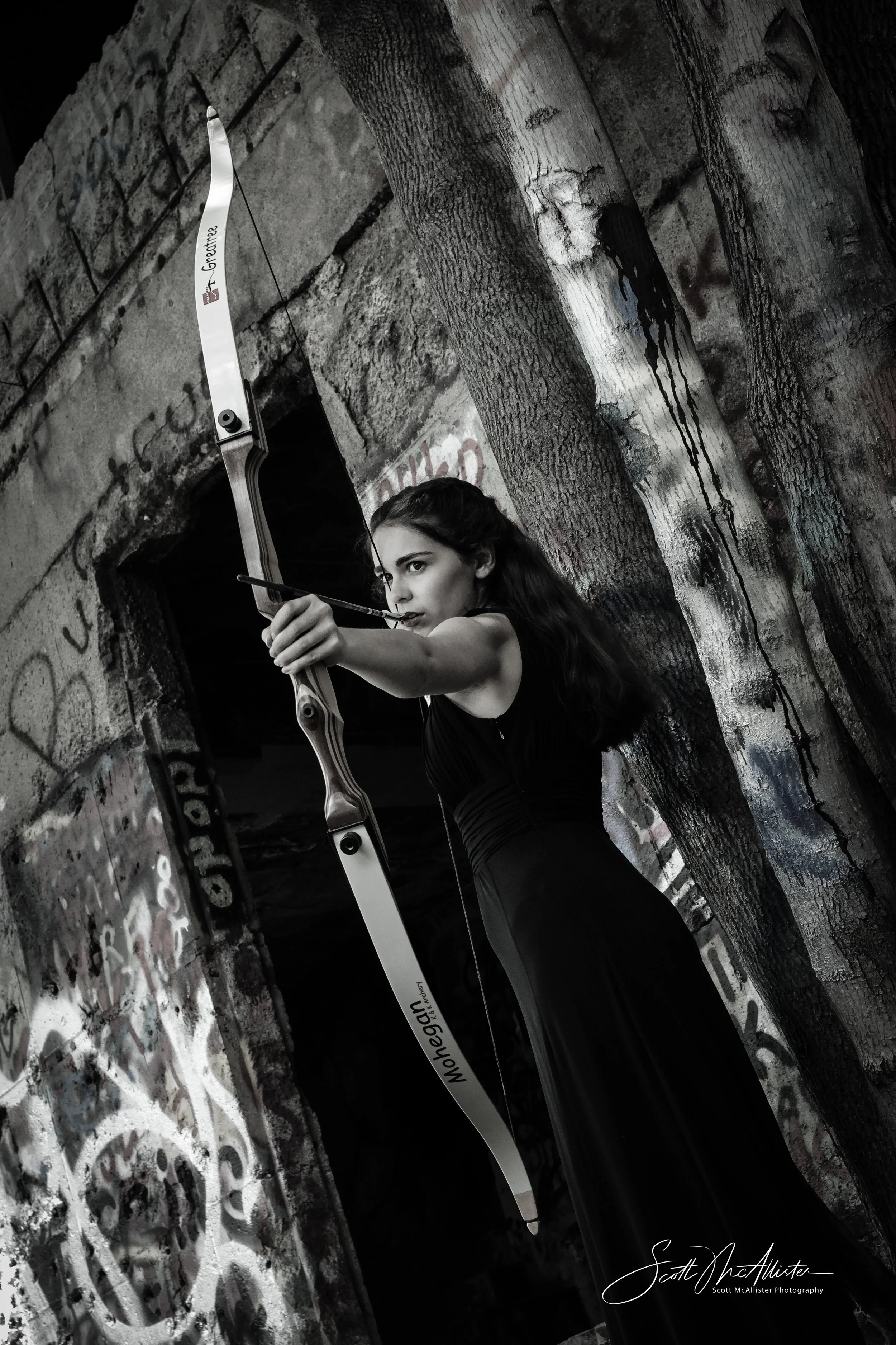 A woman aiming a bow and arrow with a determined expression in an urban, graffiti-covered setting.
