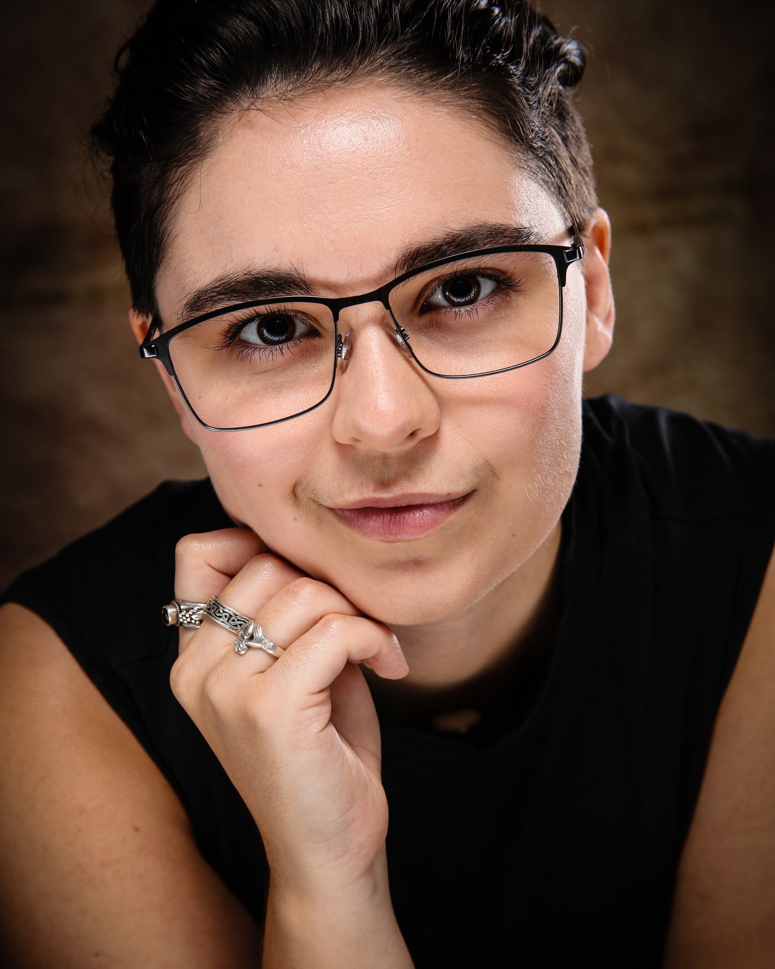 Close-up of a woman with short dark hair, wearing glasses and a black top, resting her chin on her hand with rings on her fingers, looking into the camera.