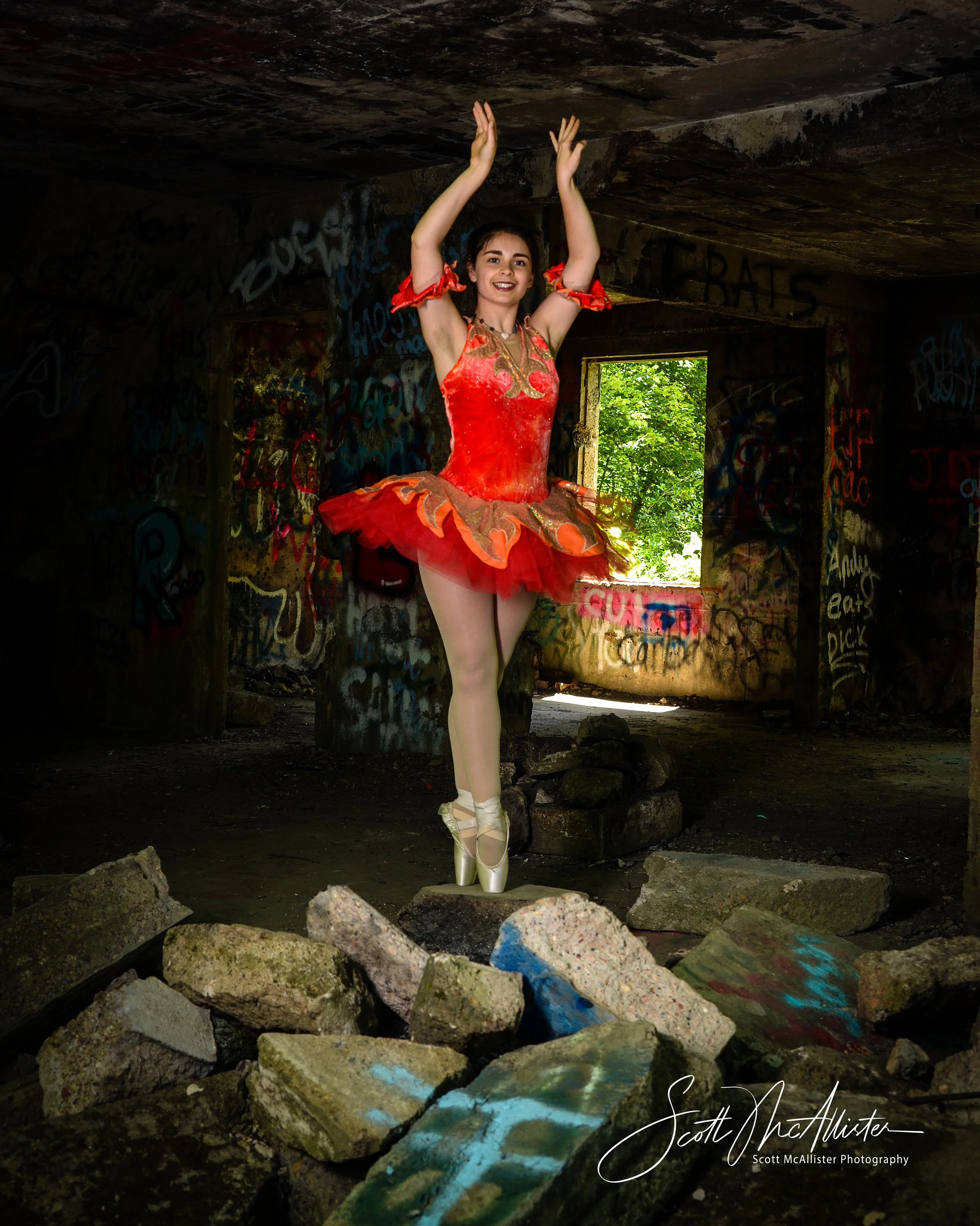 A ballerina in a red and gold costume stands on her toes in an abandoned, graffiti-covered building with natural light coming through a window, surrounded by rocks.