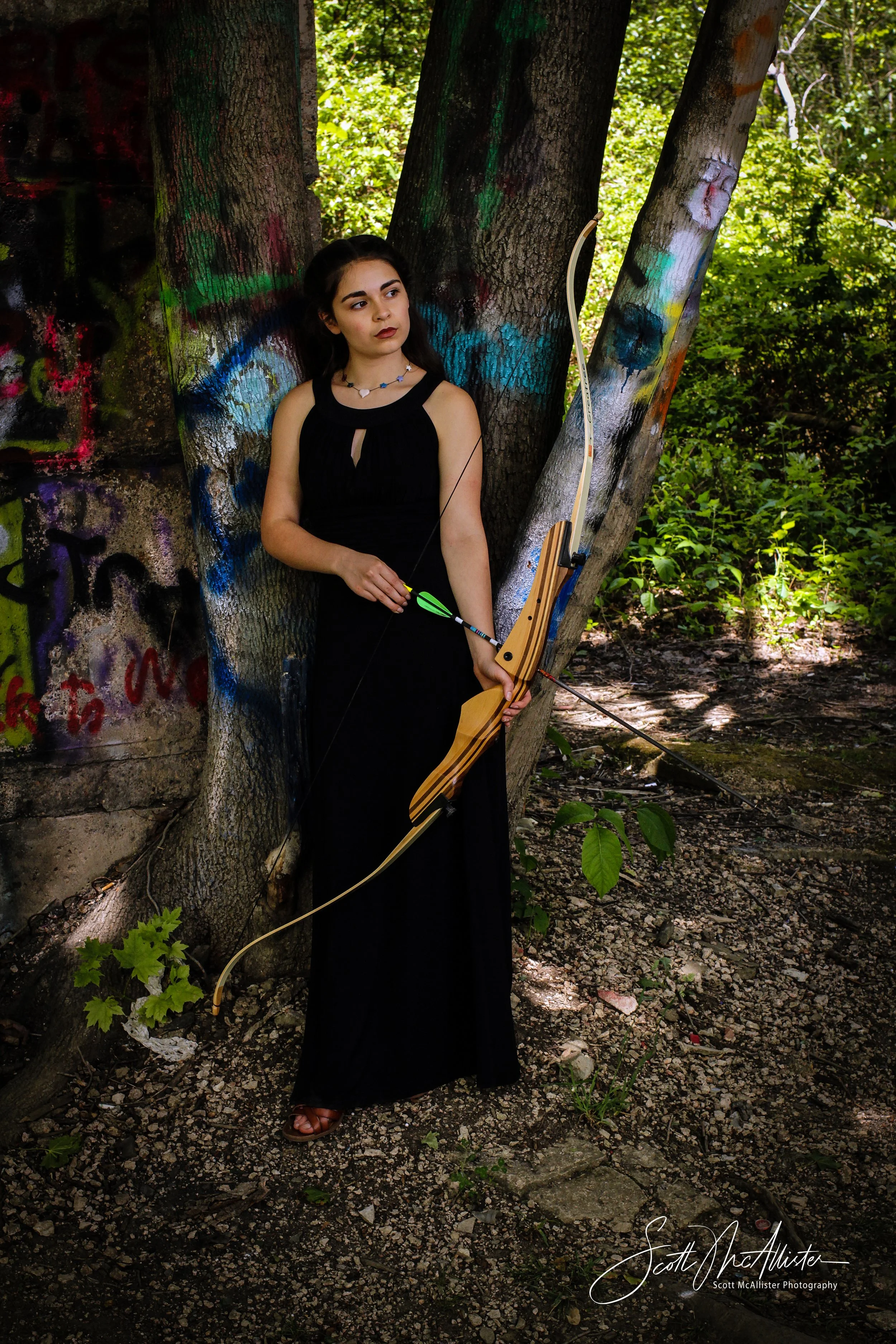 A young woman wearing a long black dress and brown sandals standing in a wooded area. She is holding a bow and arrow, with the arrow nocked and ready to shoot. The background features a tree with graffiti and a stone wall, with green foliage and sunl