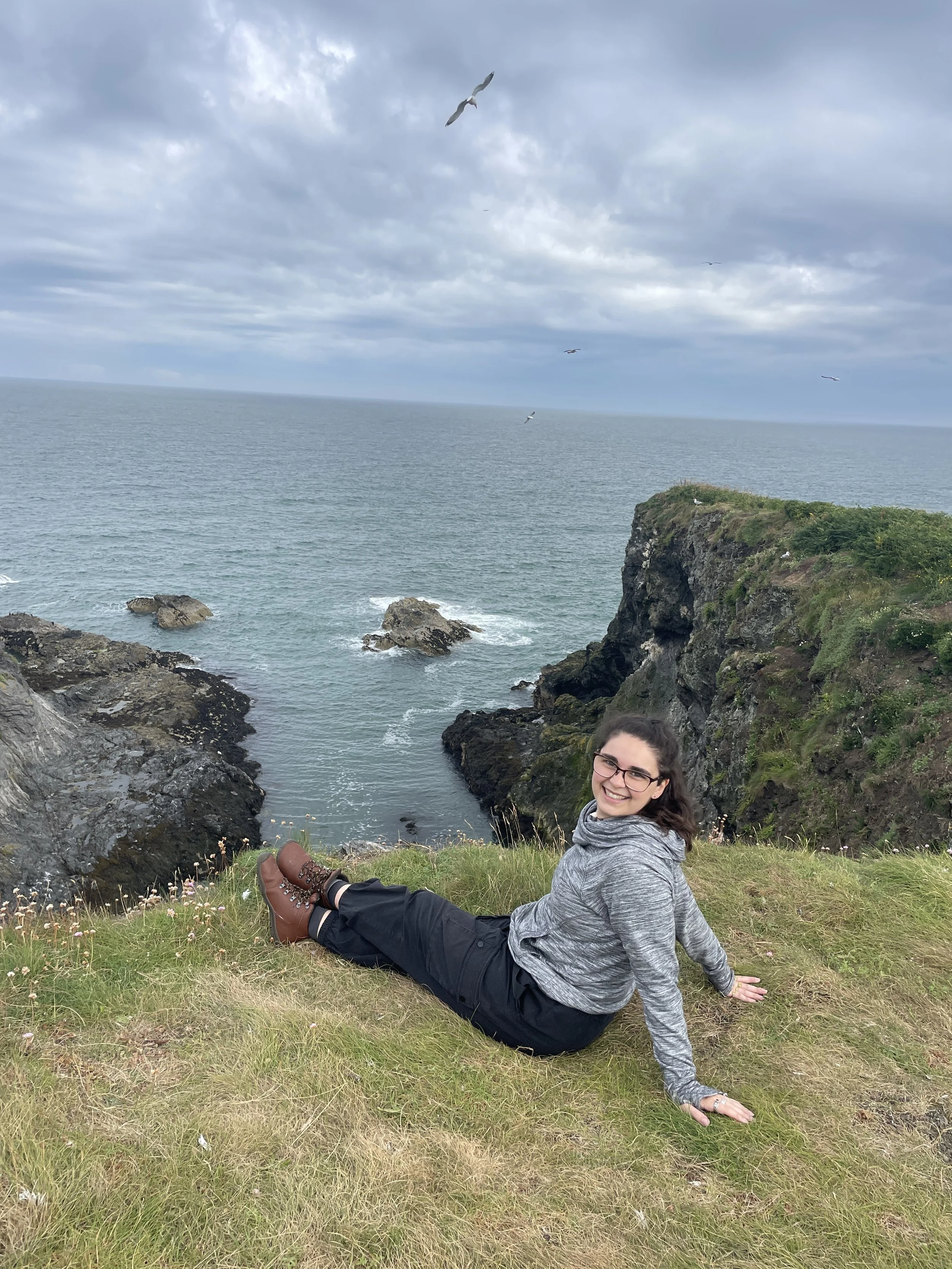 A young woman with glasses and a gray hoodie sitting on grass near a coastline with rocks, cliffs, the ocean, and a cloudy sky, smiling at the camera.