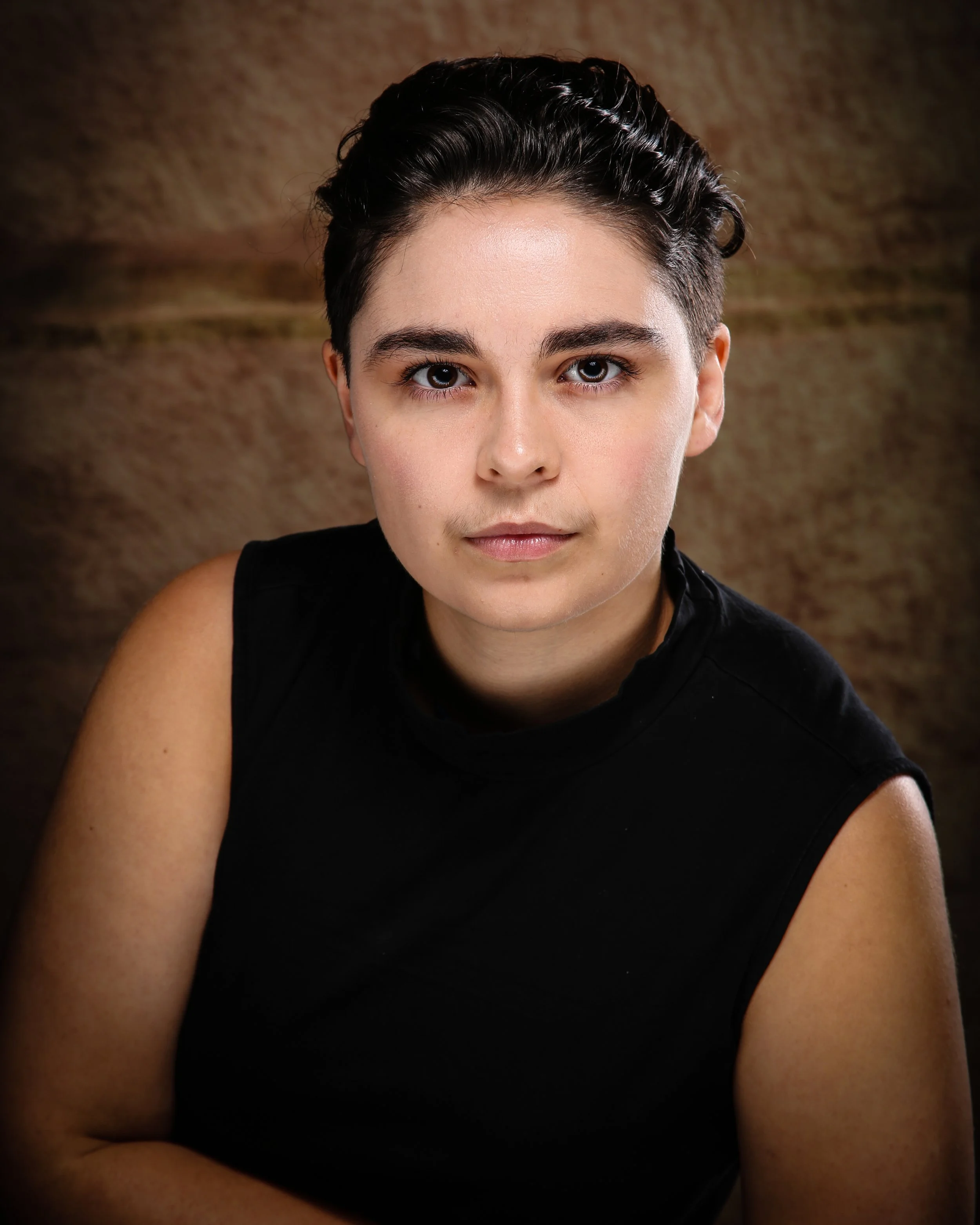Close-up of a woman with short dark curly hair, wearing a black sleeveless top, posing in front of a brown textured background.