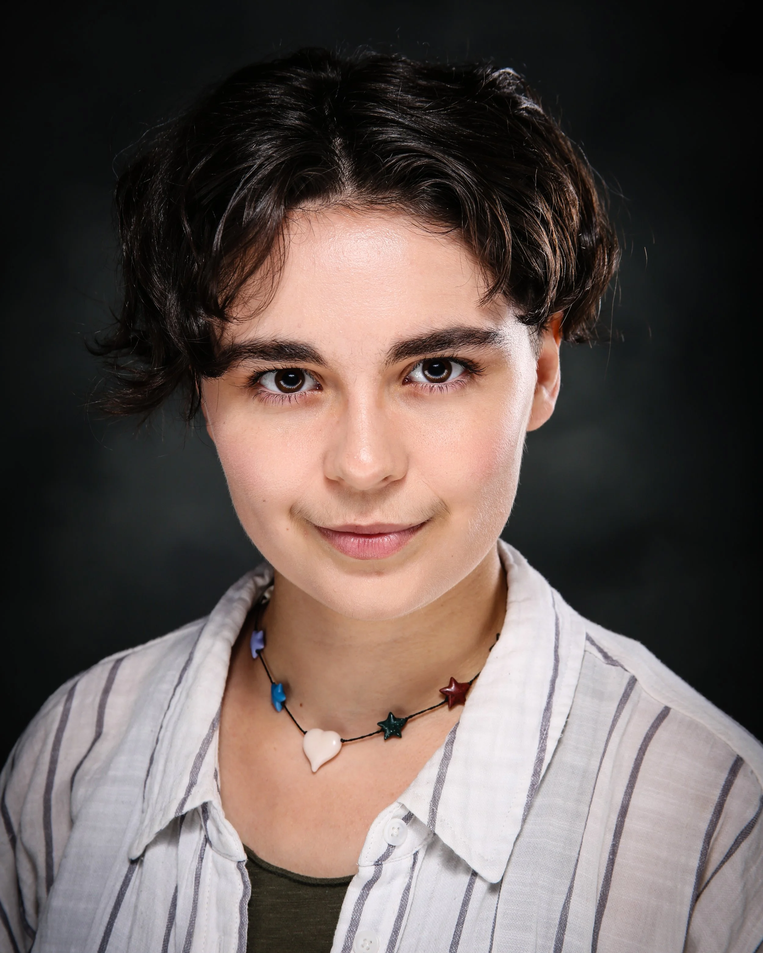 A person with short, dark, curly hair, wearing a striped shirt and a black beaded necklace with star-shaped and heart-shaped charms, smiling softly against a black background.