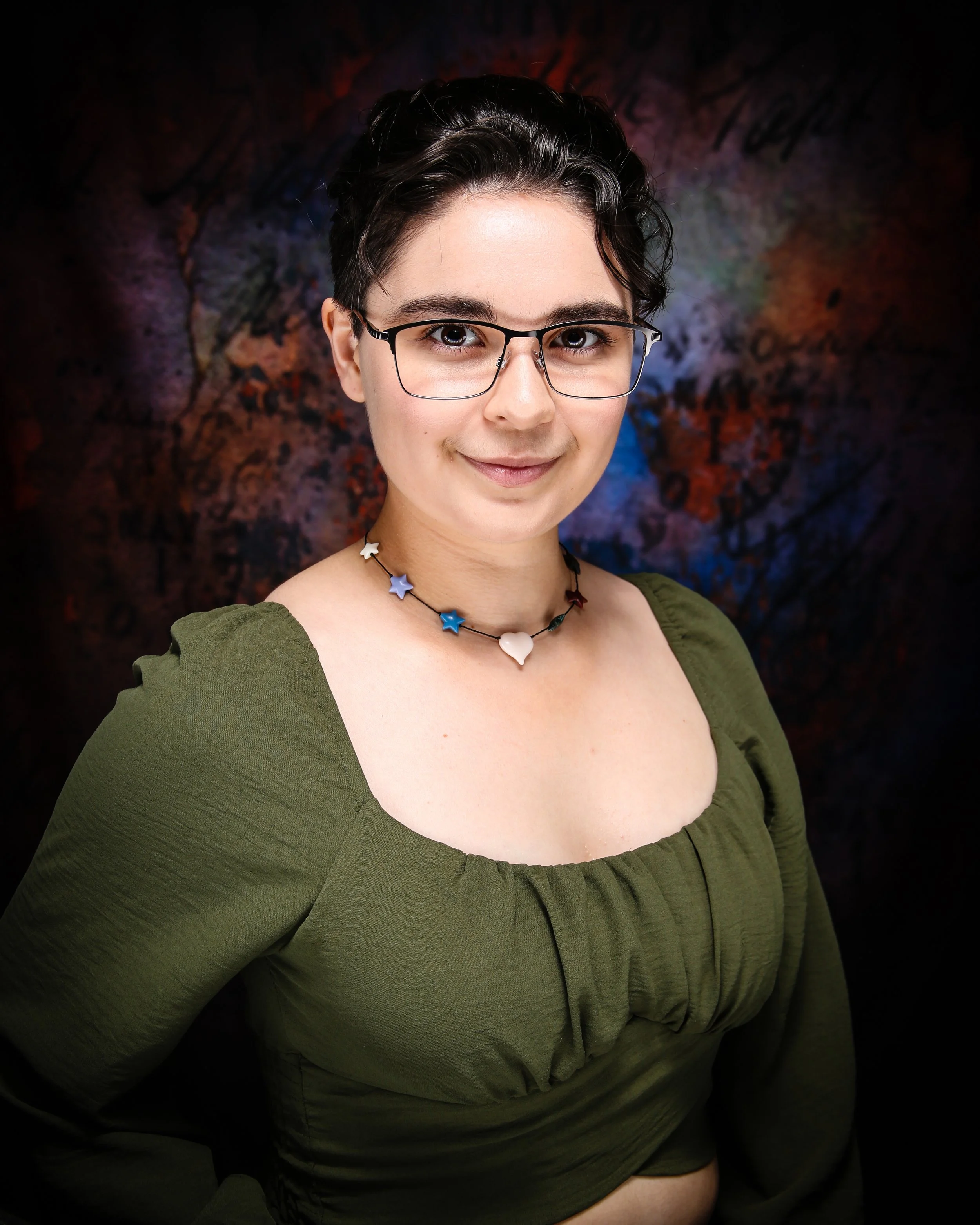 Close-up of a smiling woman with short dark hair, glasses, and star-shaped necklace, wearing a green top, against a colorful abstract background.