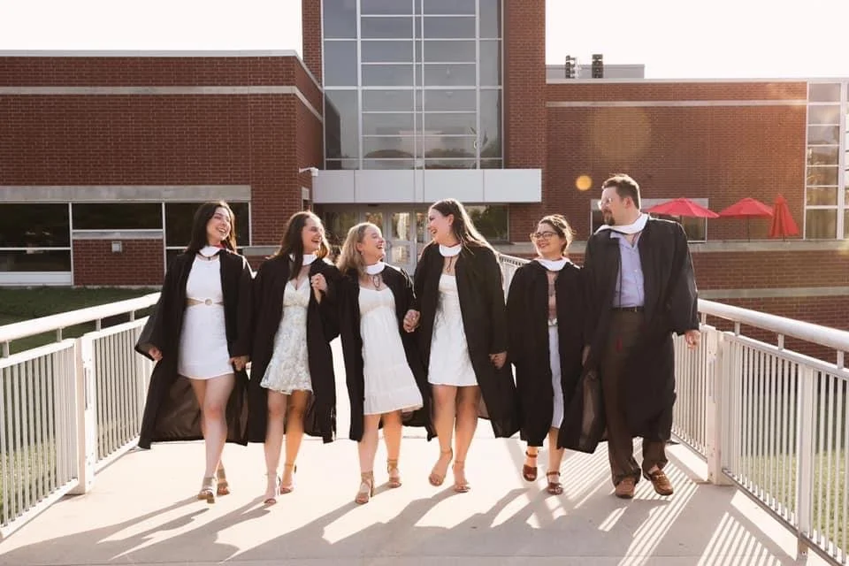 Graduates in caps and gowns walking on a bridge outside a brick and glass building, smiling and holding hands.
