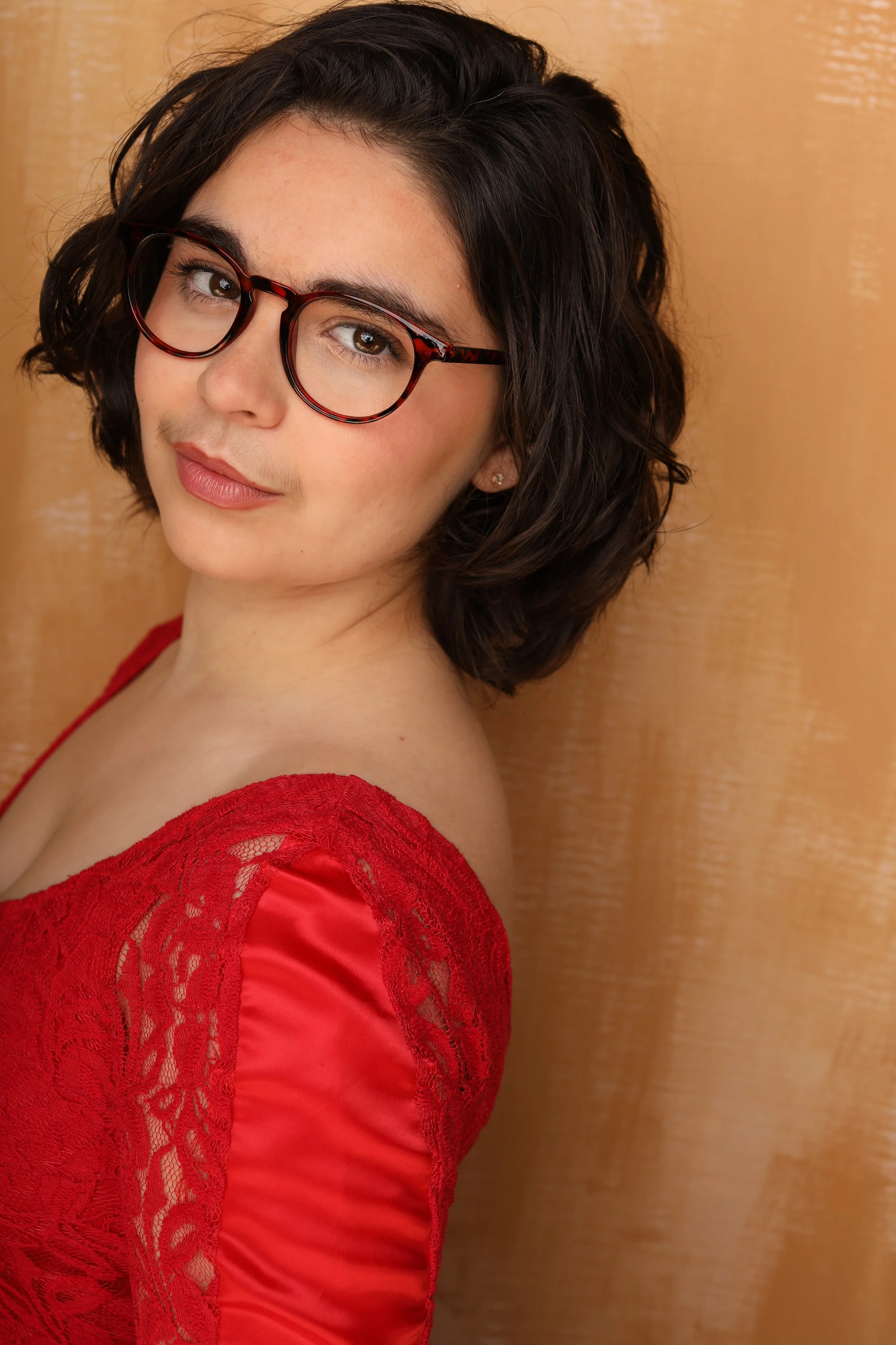 Close-up of a woman with short dark wavy hair wearing red lace dress and red eyeglasses with tortoise pattern, leaning against a beige textured wall.