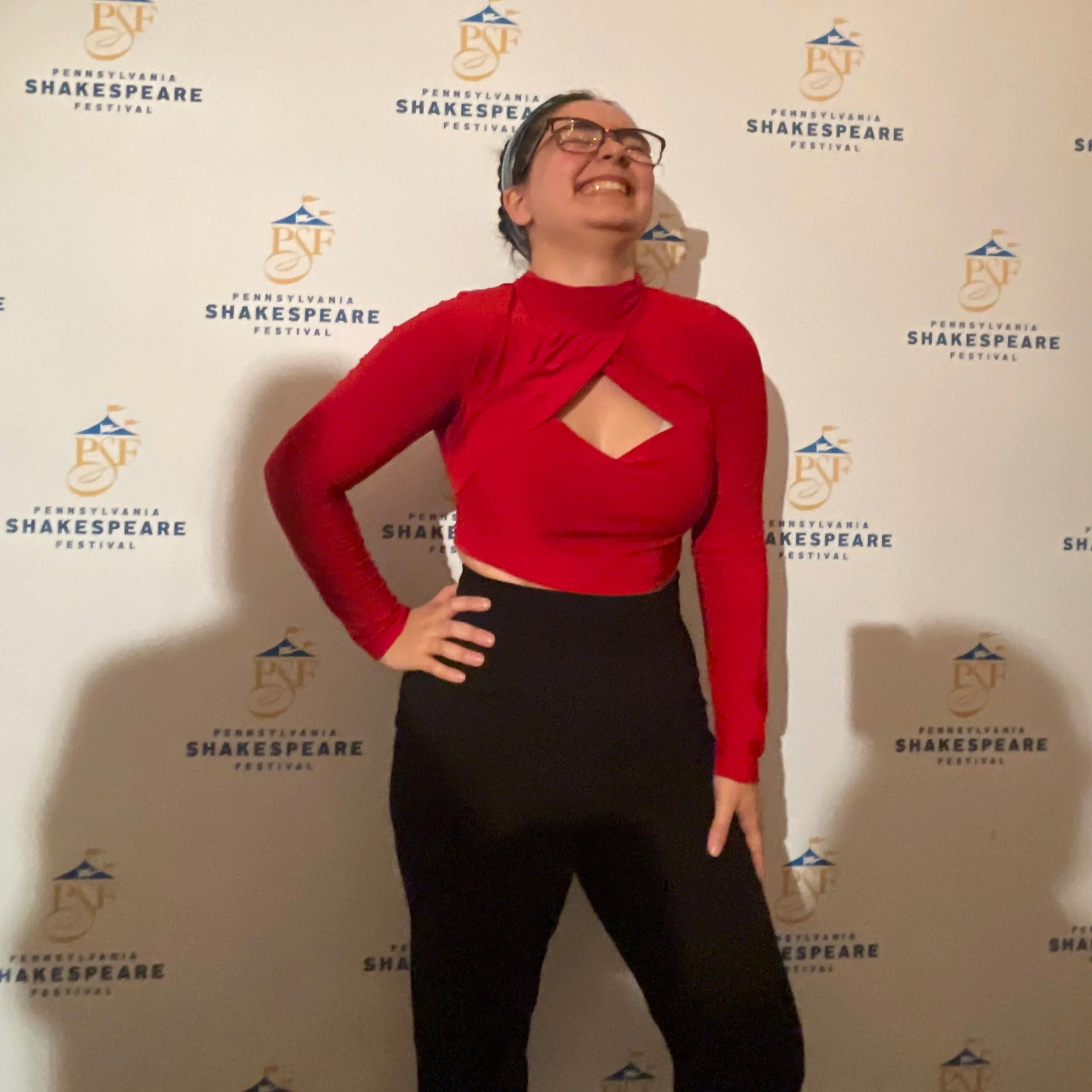 A woman in a red top and black pants posing with one hand on her hip and smiling at the camera in front of a step and repeat banner with the Pennsylvania Shakespeare Festival logo.