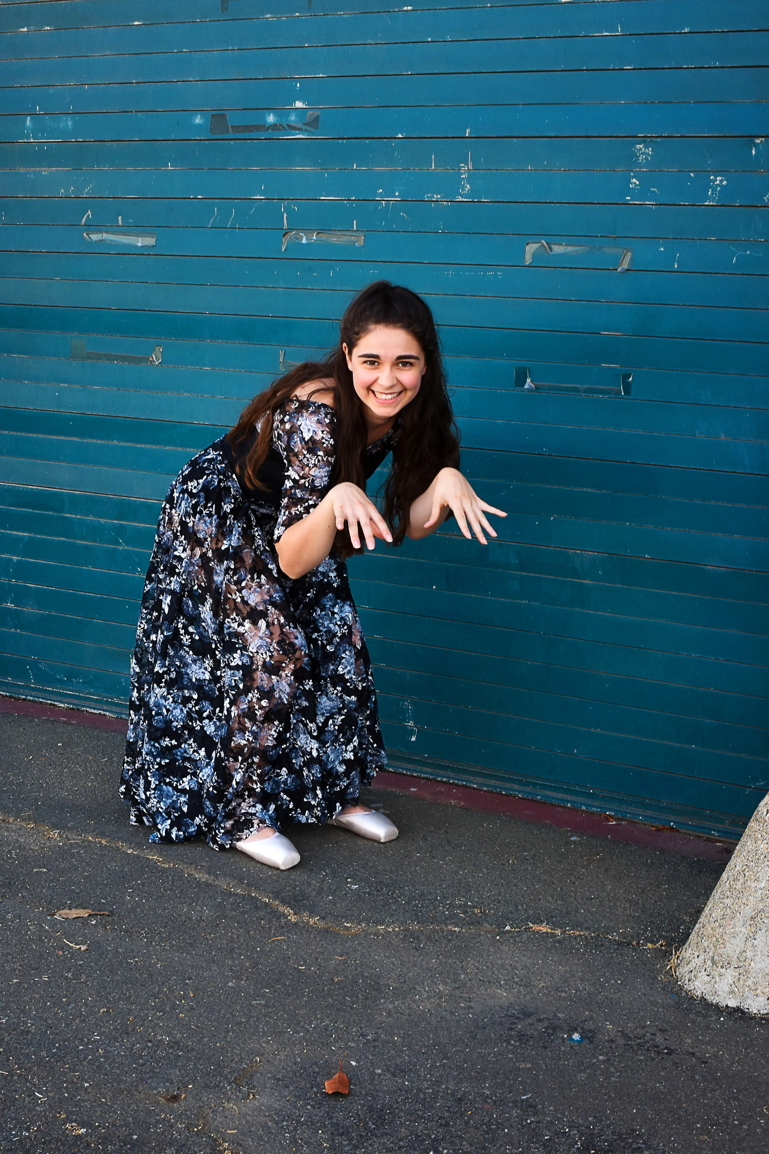 A young woman with long dark hair, wearing a black dress with a floral pattern and white ballet flats, is smiling and crouching in front of a blue metal garage door with horizontal panels. She has her hands raised, mimicking claws or paws, with a pla