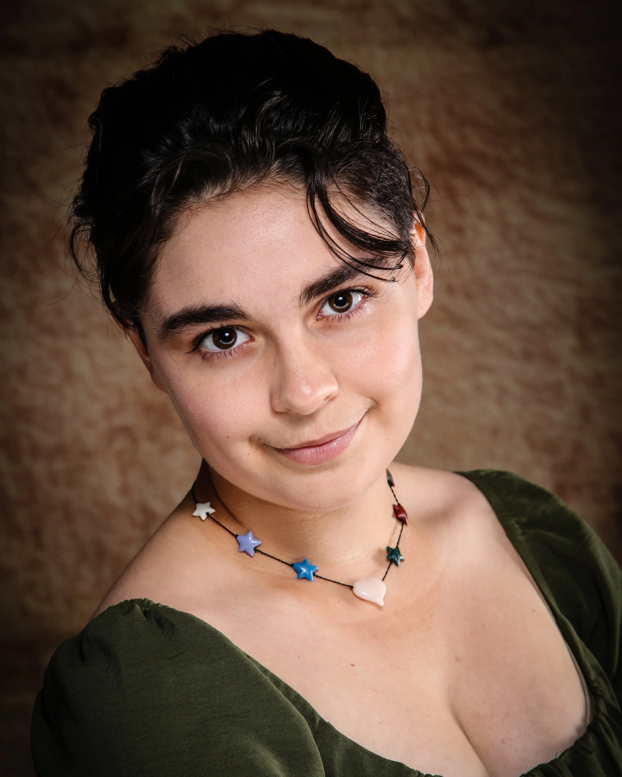 Close-up of a woman with short black hair, wearing colorful star-shaped and shell necklaces, and an off-shoulder green top, smiling softly against a textured brown background.