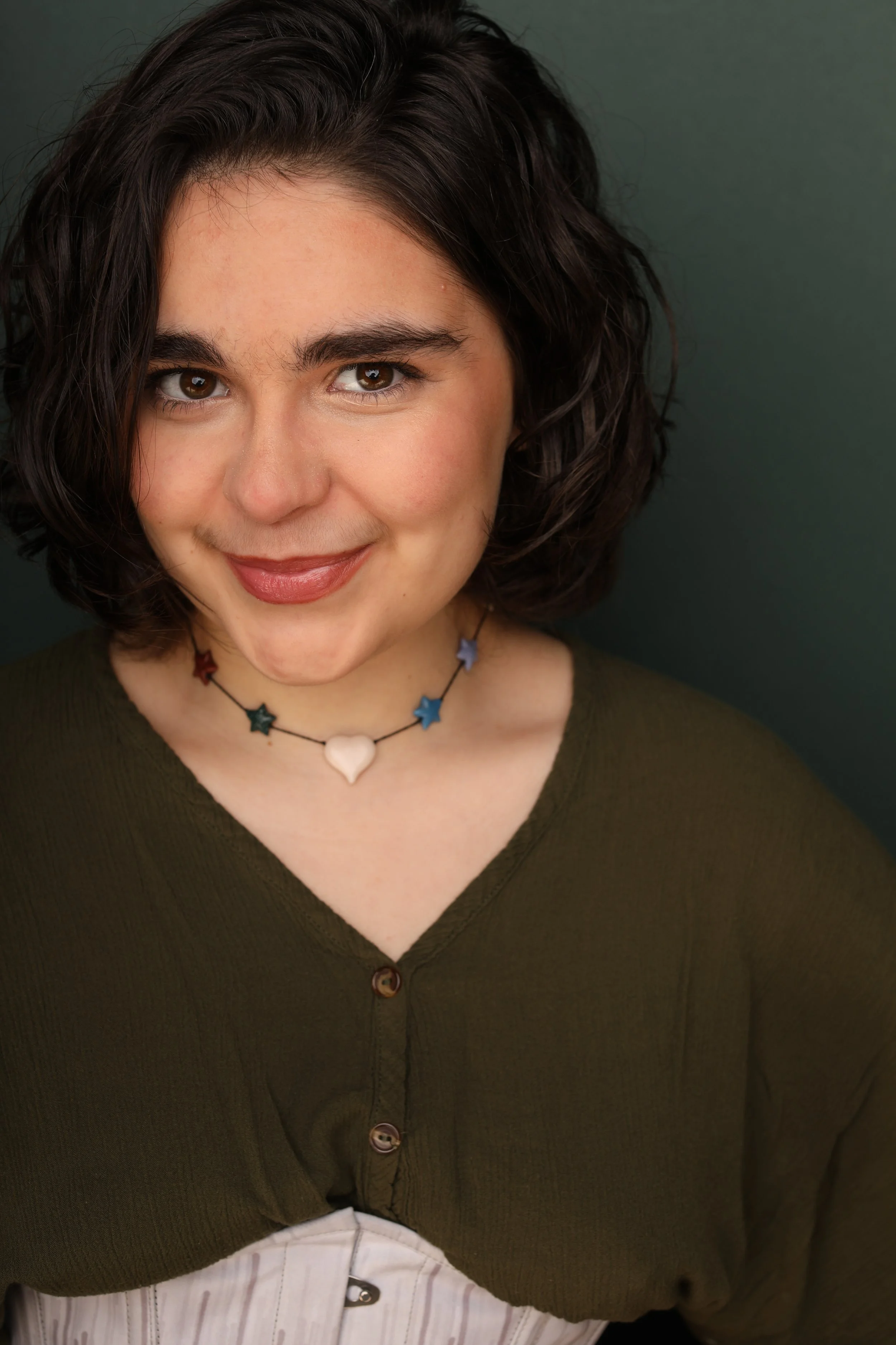 A woman with short, dark, wavy hair and brown eyes, wearing a dark green button-up shirt and a star and heart necklace, smiling slightly, against a dark background.