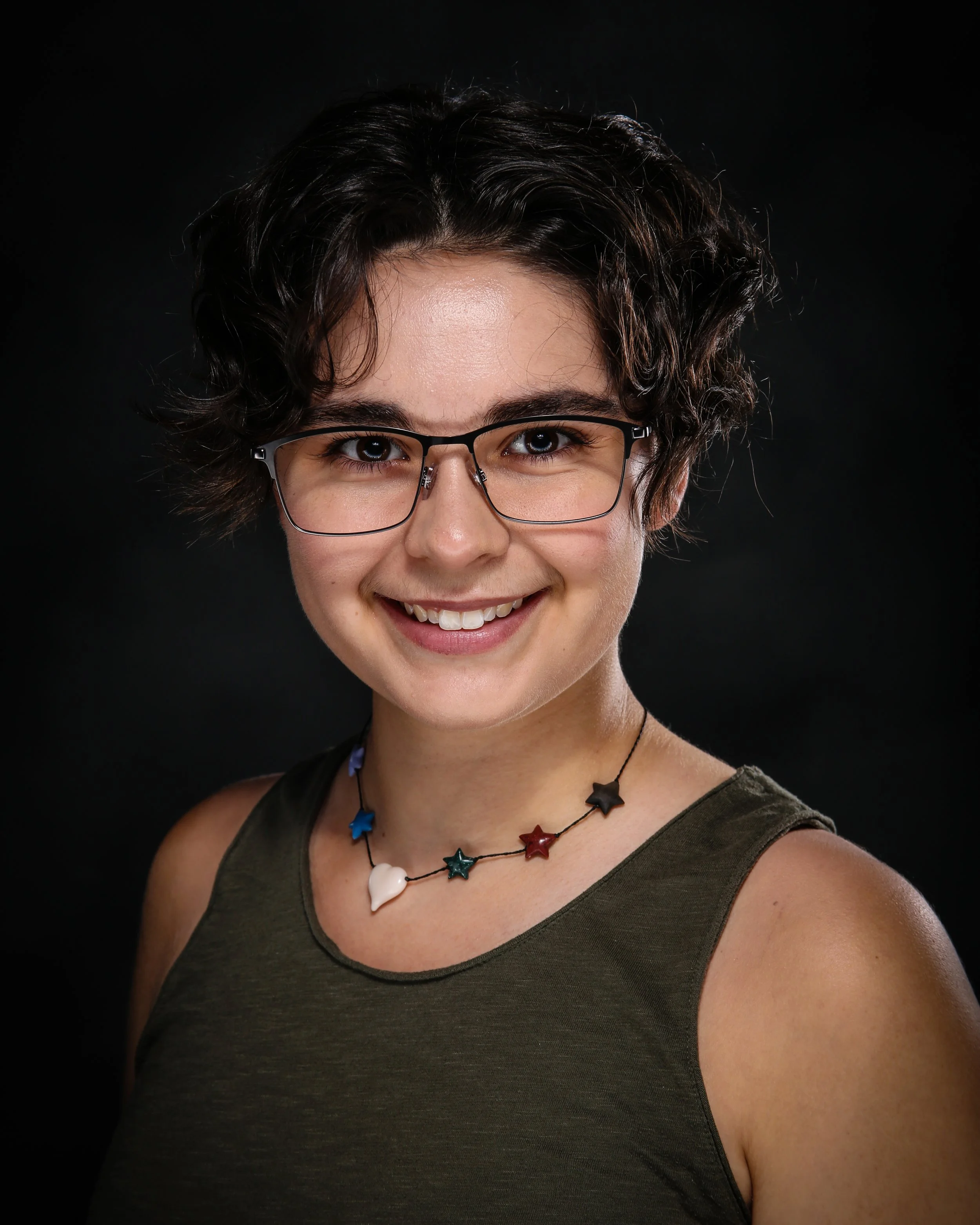 Close-up portrait of a smiling woman with short, curly dark hair, wearing glasses, a dark green sleeveless top, and colorful star and heart-shaped necklace against a black background.