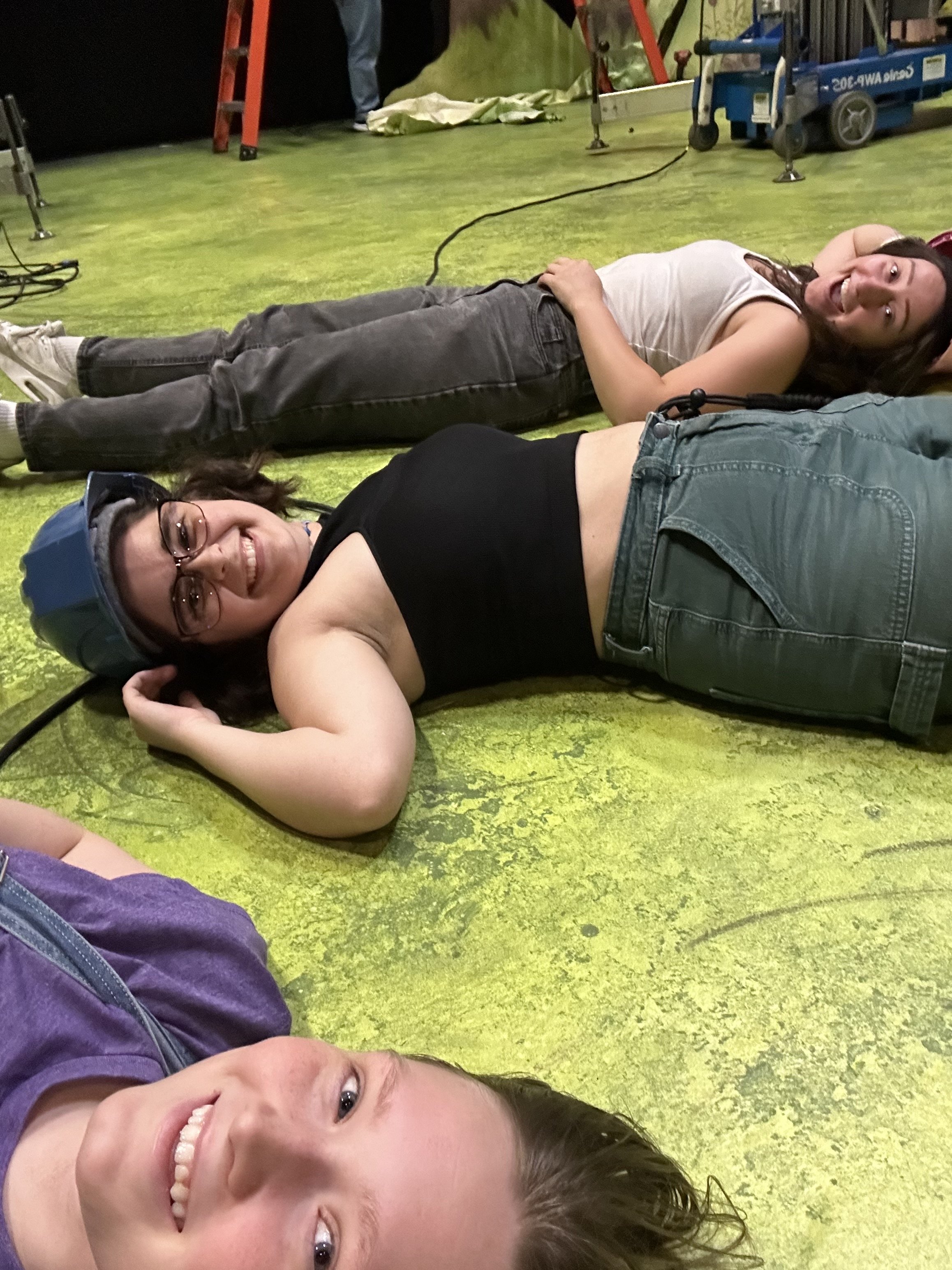 Three smiling young women lying on a green carpeted floor, taking a selfie with one in the foreground and the other two in the background.