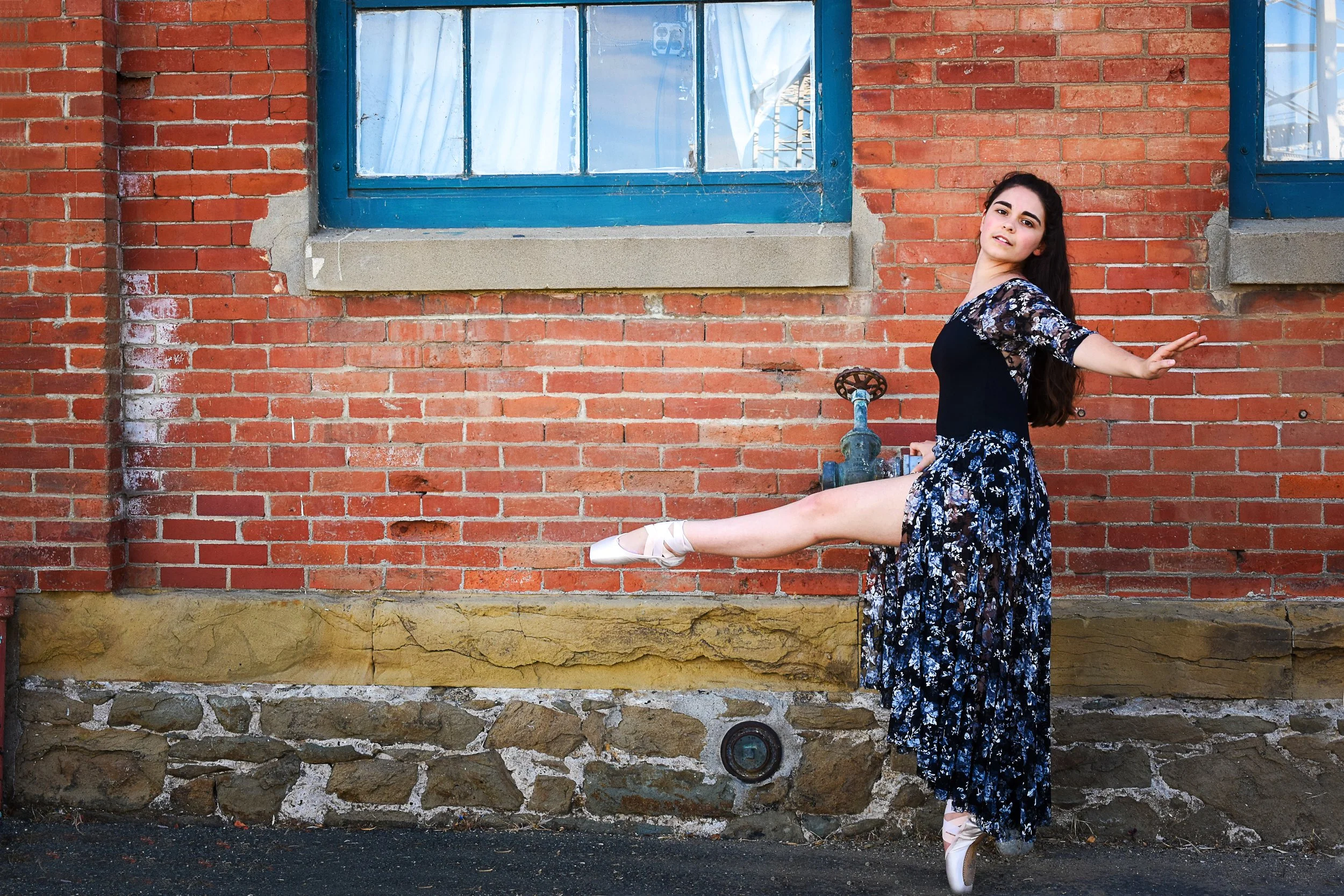 A woman in a floral dress and ballet pointe shoes poses with her right leg extended, against a red brick wall and window with blue frames, near an old outdoor water faucet.