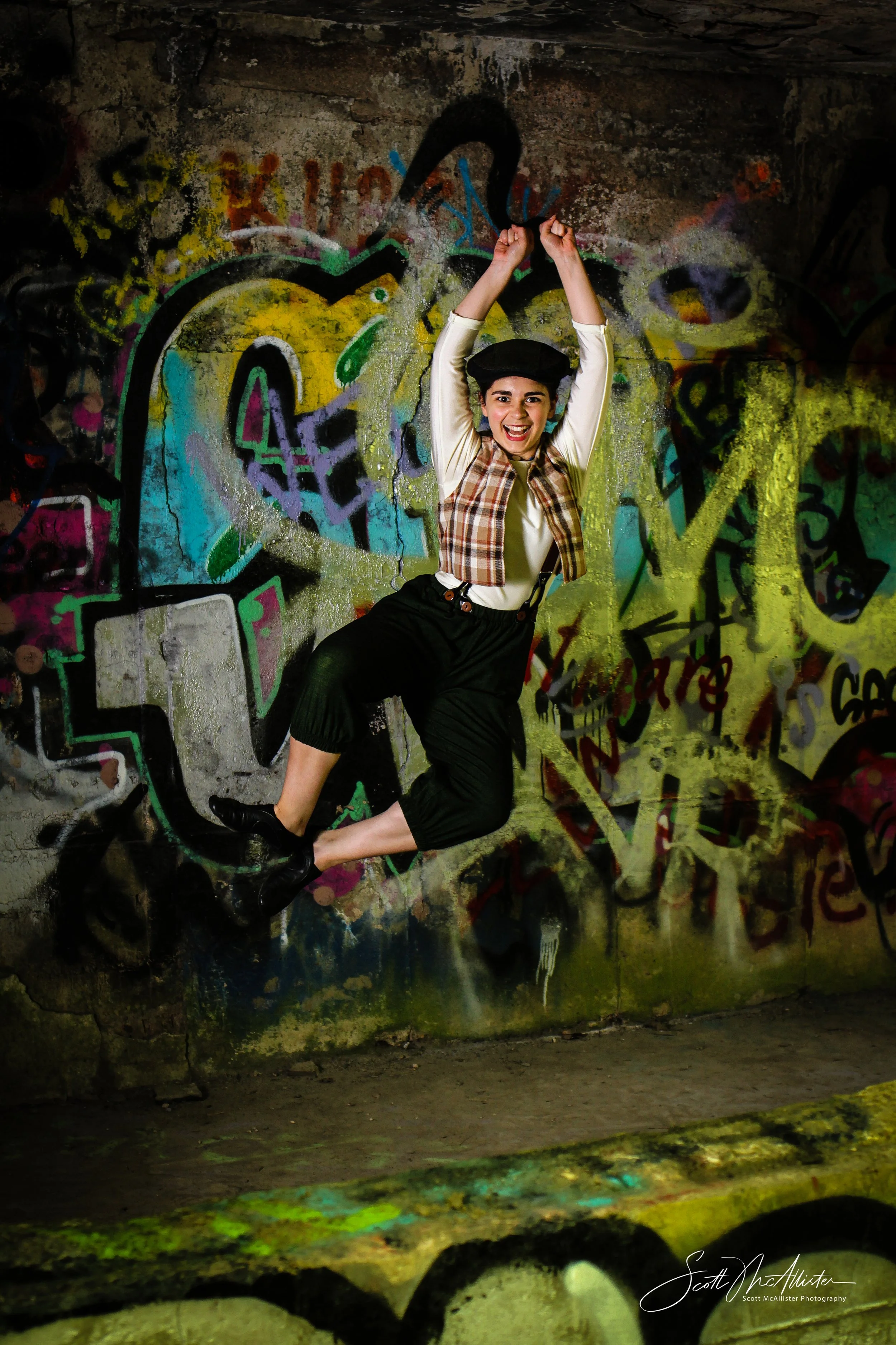A young woman in a plaid vest and black shorts joyfully poses with arms raised in front of a colorful graffiti wall. She is wearing a black beret and is standing in a dimly lit space with a concrete floor.
