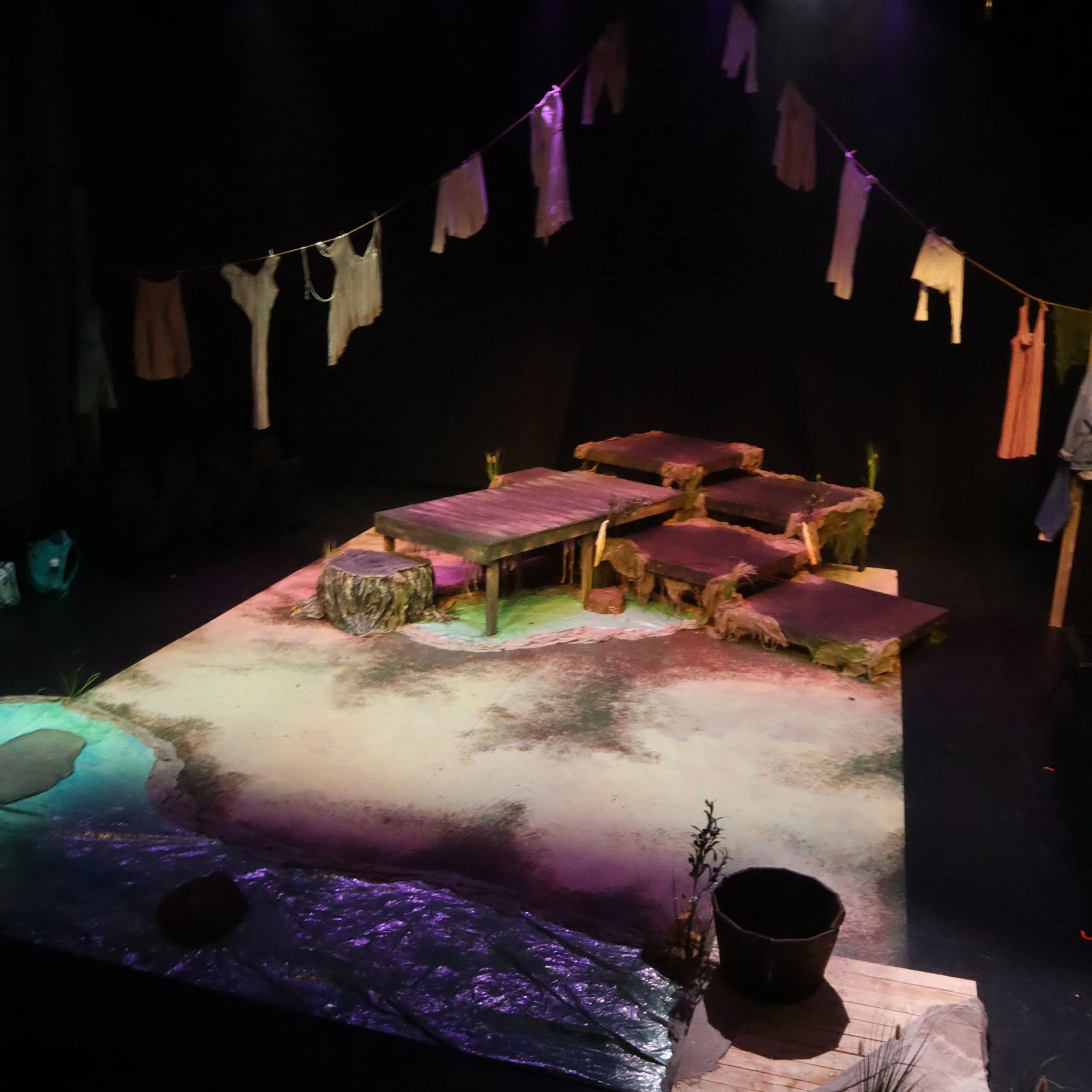 A nighttime scene with floating pieces of fabric or paper hung on a line, illuminated by purple and pink lighting, over a body of water with rocks and potted plants in the foreground.