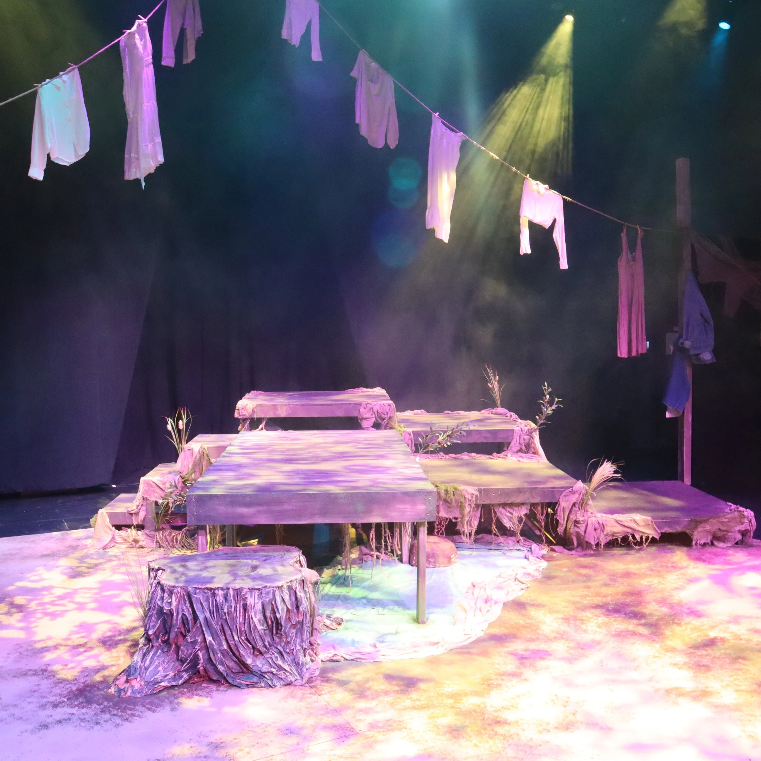 Stage set with hanging laundry on a line, illuminated by colorful stage lights, with layered platforms and surrounding plants.