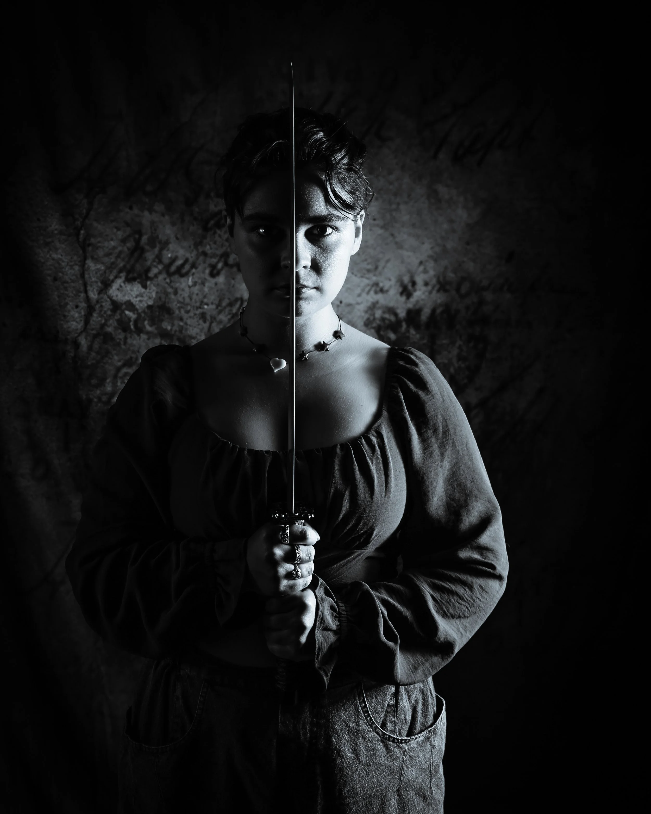 A young person with short hair, holding a sword vertically in front of their face. The person is wearing a necklace and a dark top. The photo is in black and white, with a dark background.