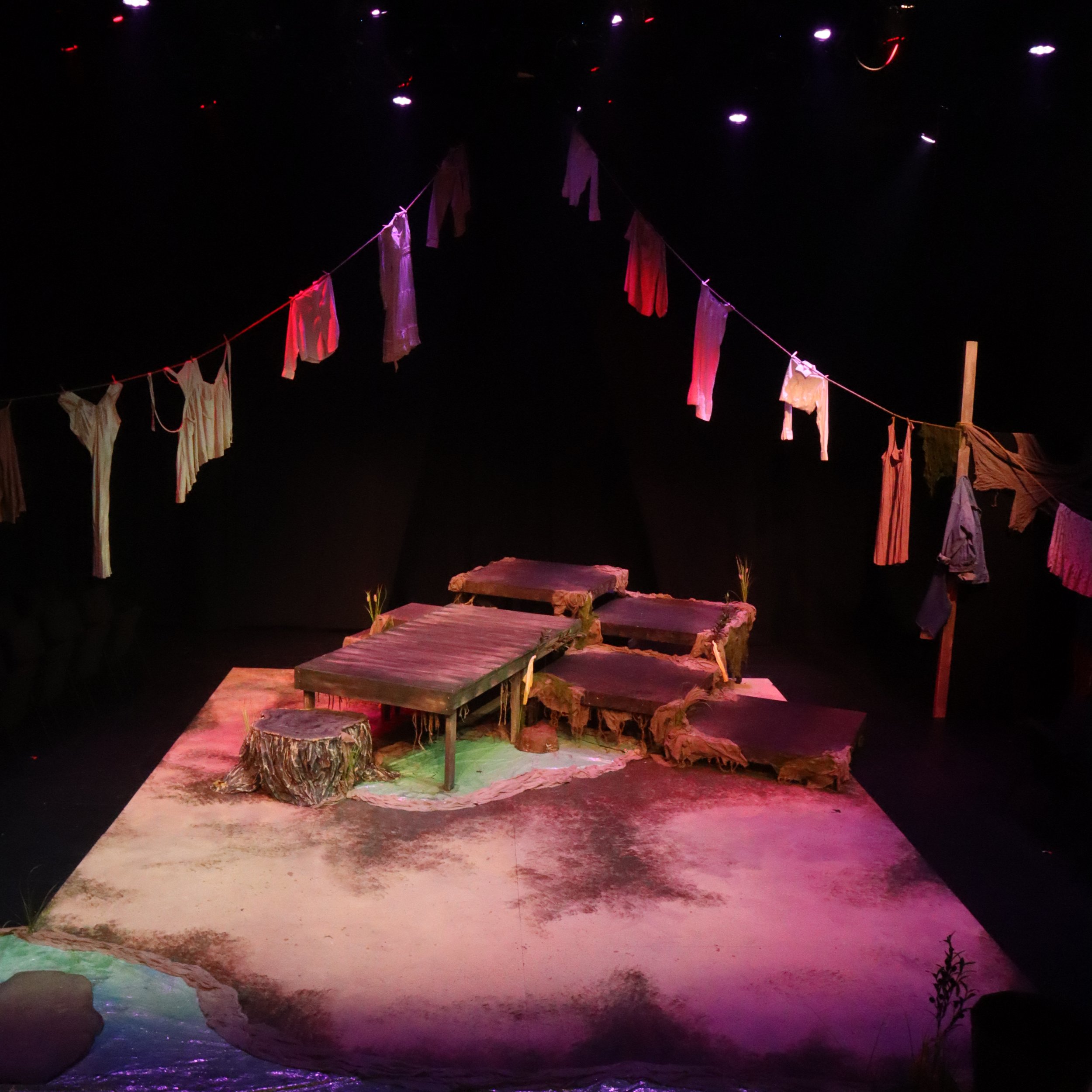 Stage set with hanging clothes, wooden planks, and a water feature, illuminated with colored lights in a theatrical or performance setting.