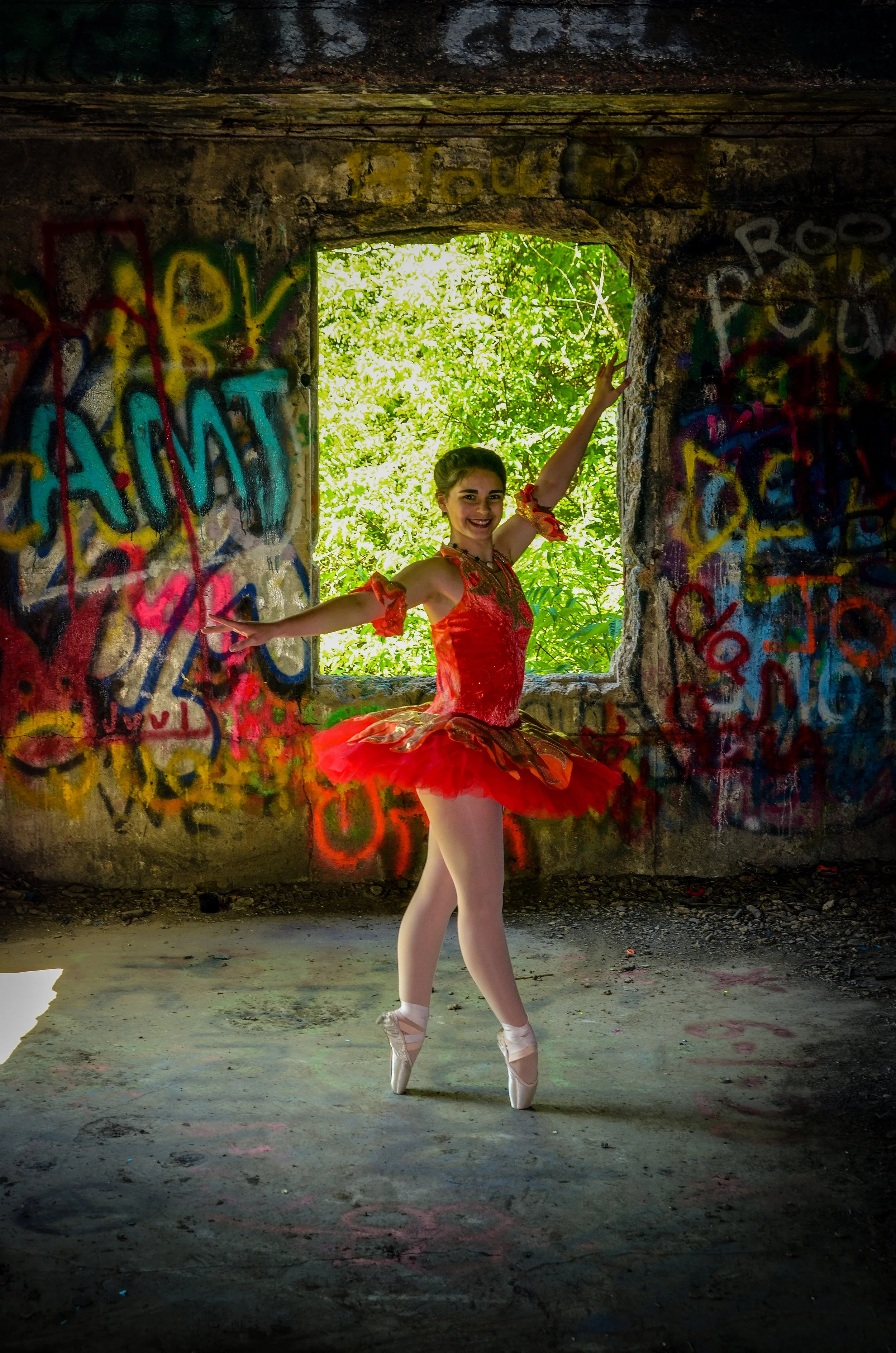 A young woman in a red ballet costume and pointe shoes dancing inside an abandoned, graffiti-covered building with a window showing green foliage outside.