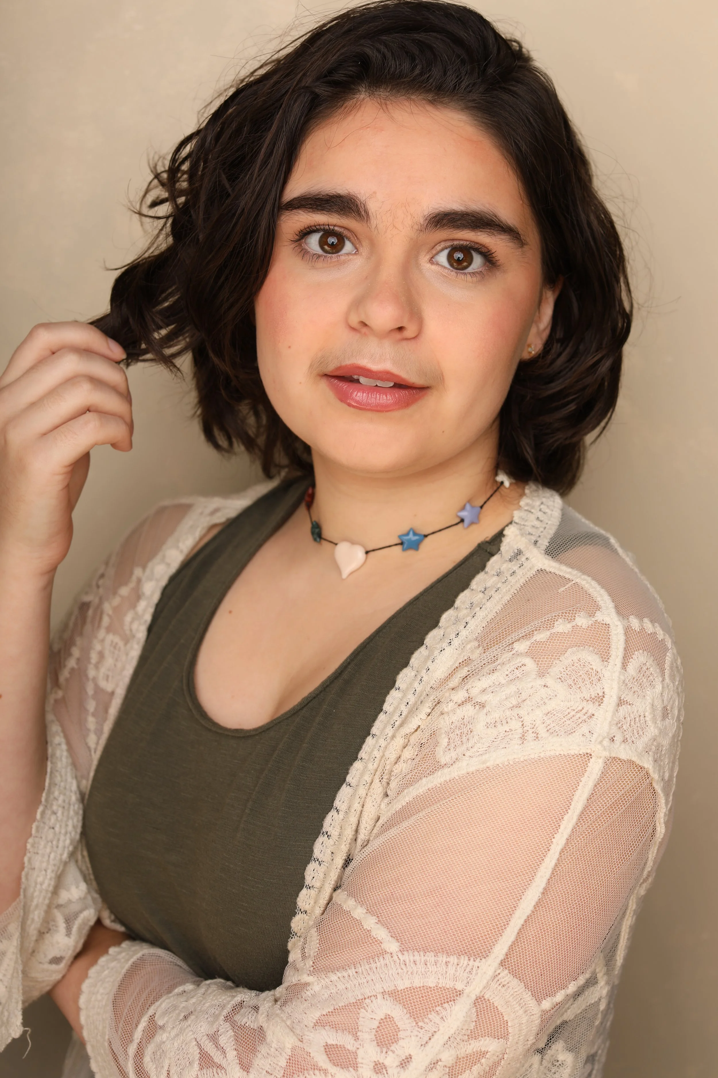 A young woman with short dark brown hair and brown eyes, wearing a green tank top under a gray jacket and a white lace cardigan. She has a necklace with star and heart charms and is touching her hair with her right hand. She has a friendly expression
