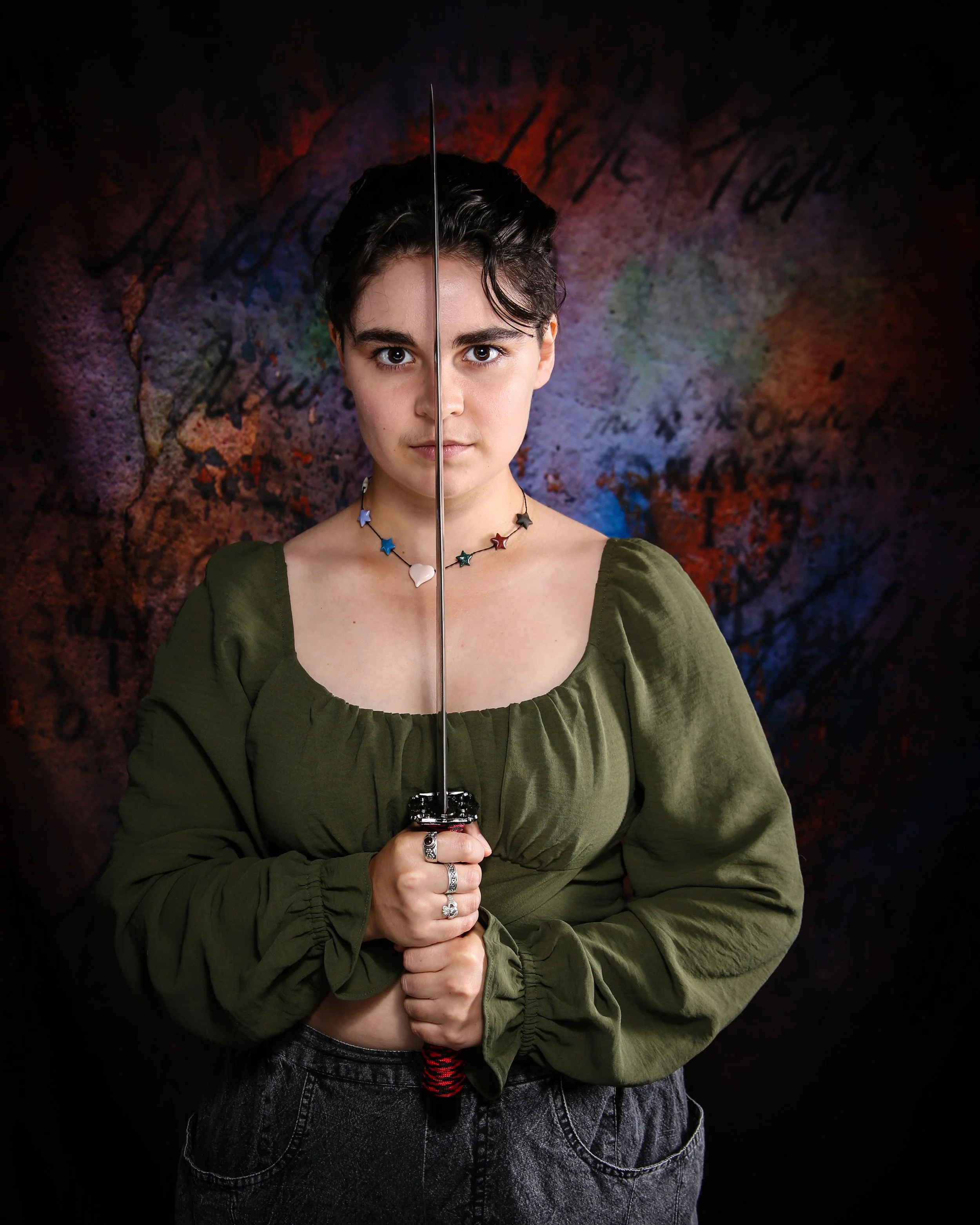 Person with short dark hair, holding a fencing sword with both hands, wearing a green top and black pants, against a colorful, textured background.