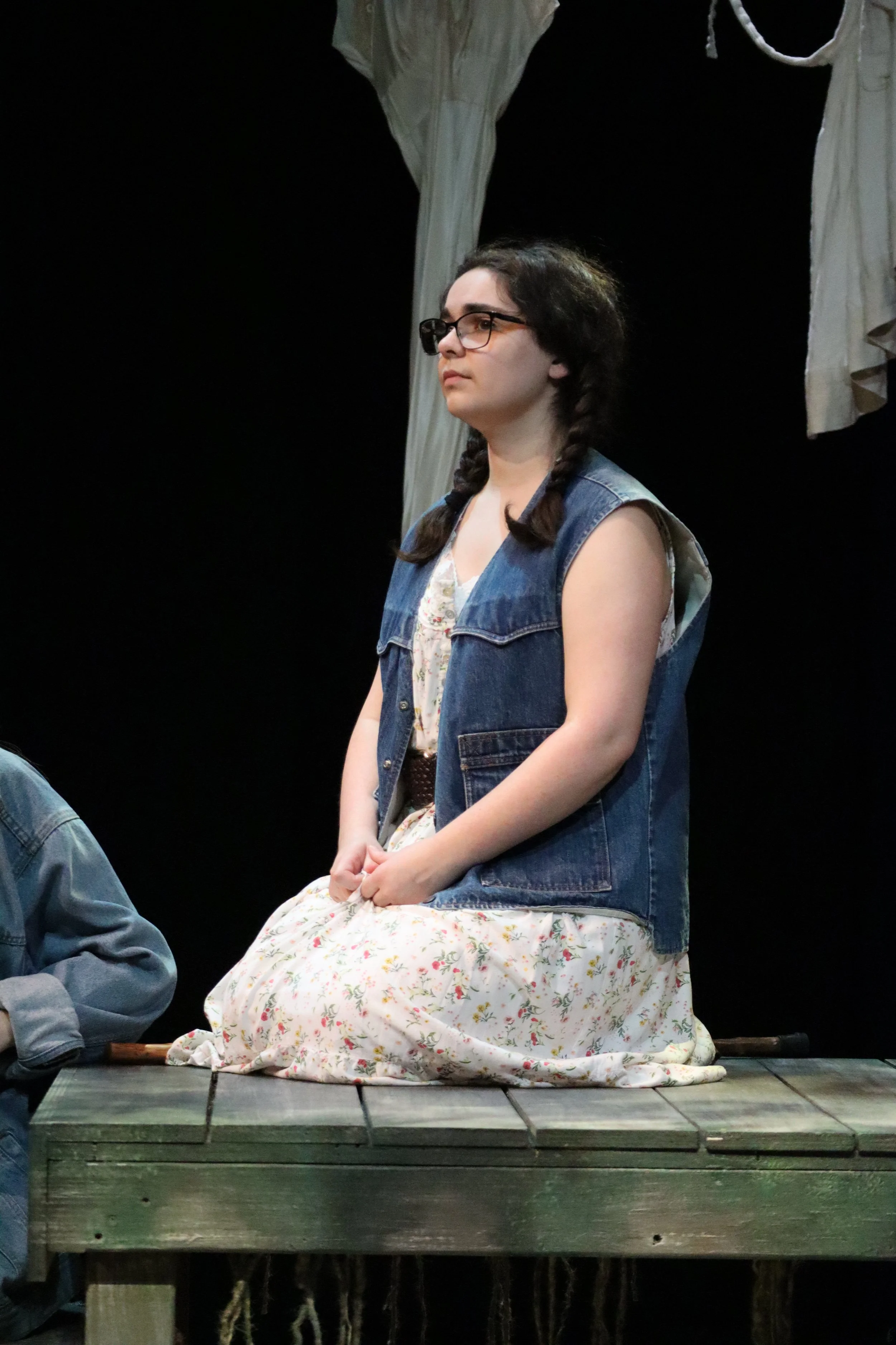 A person sitting on a wooden platform wearing a floral dress and a denim vest, with braided hair and glasses, with fabric hanging behind them in a dark setting.